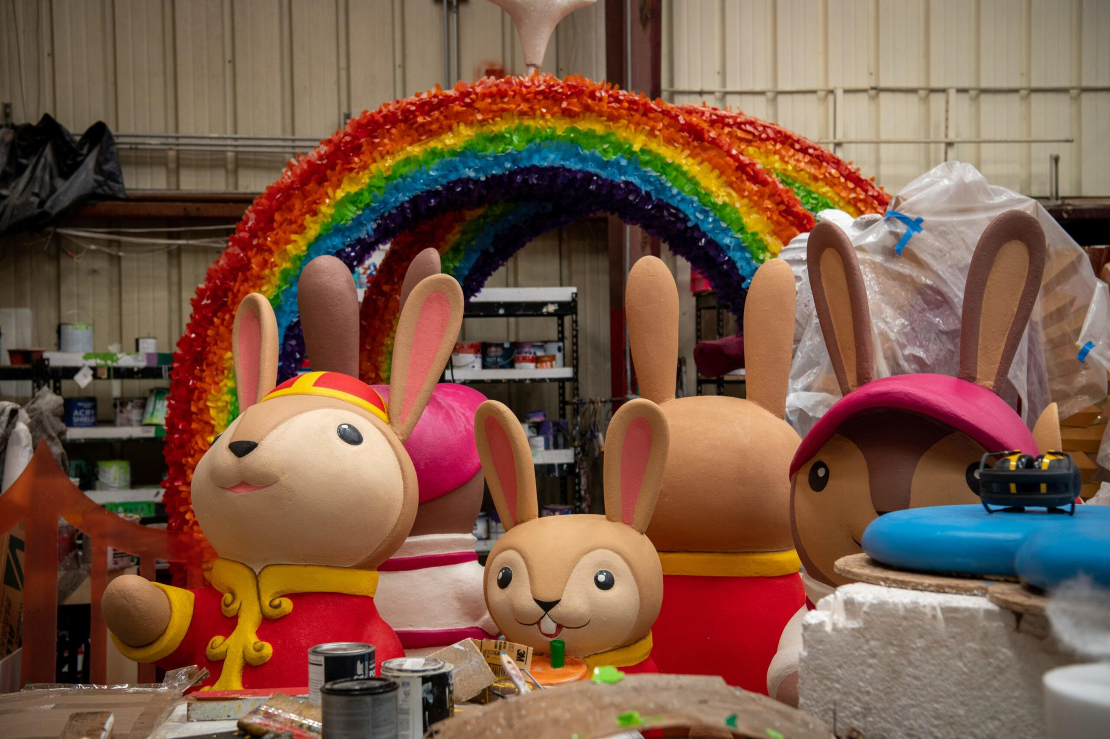 Five large rabbit figures dressed in colorful clothing are in a workshop, with a vibrant rainbow arch behind them and various art supplies scattered around.