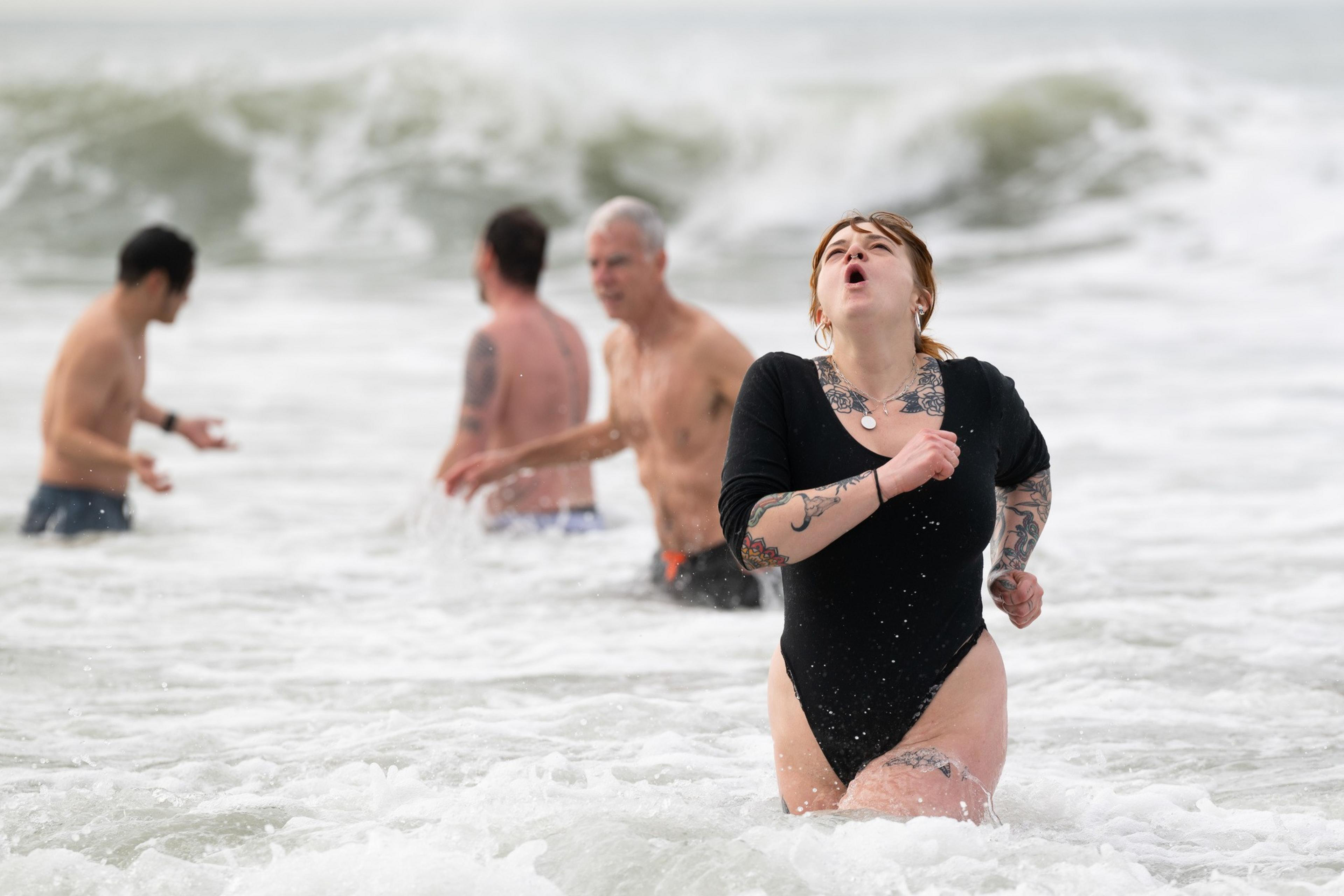 A woman in a black swimsuit, with tattoos on her arms, runs through the ocean water, while three men in the background enjoy the waves under a cloudy sky.