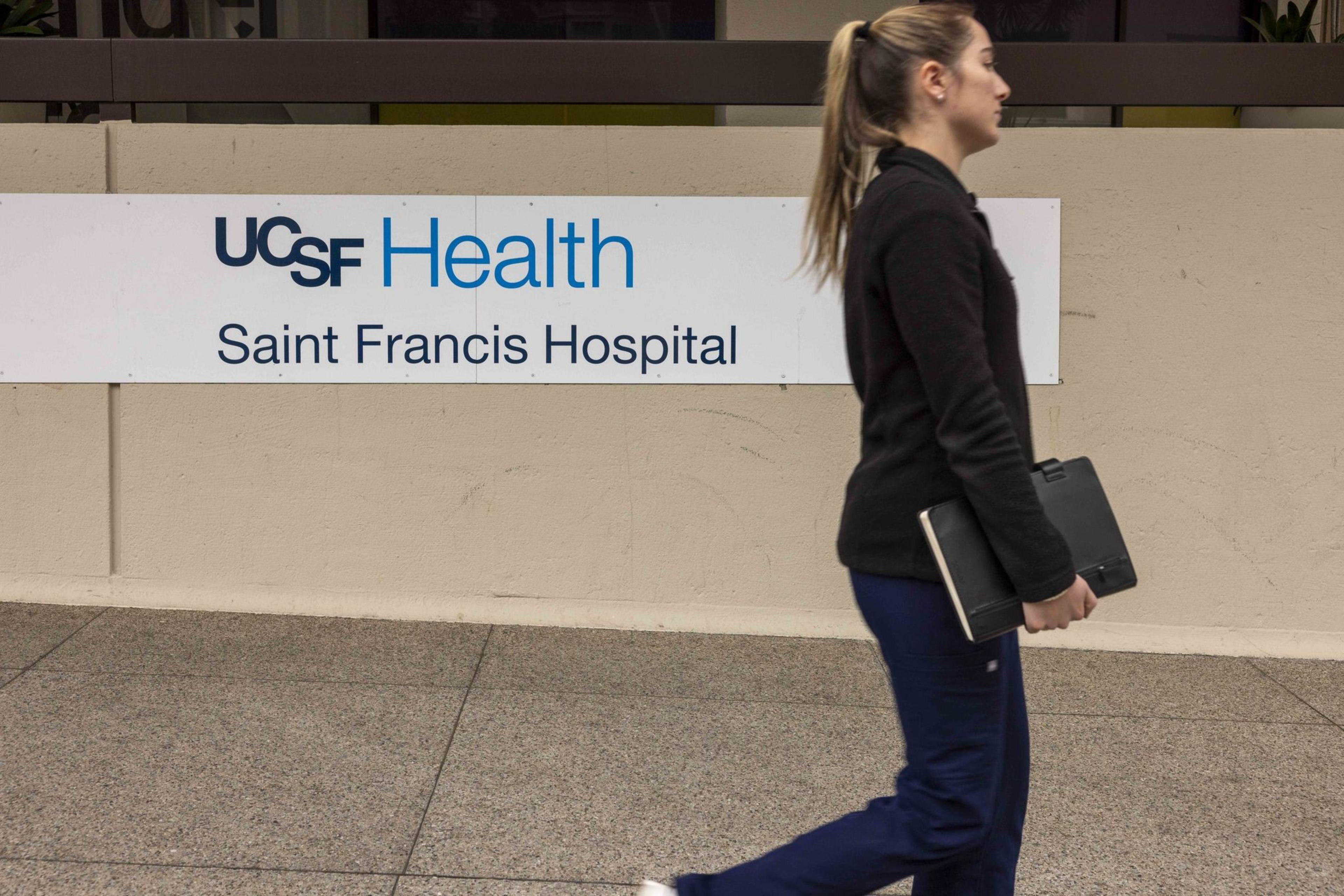 A woman is walking past a sign for UCSF Health Saint Francis Hospital, holding a closed laptop against her side.