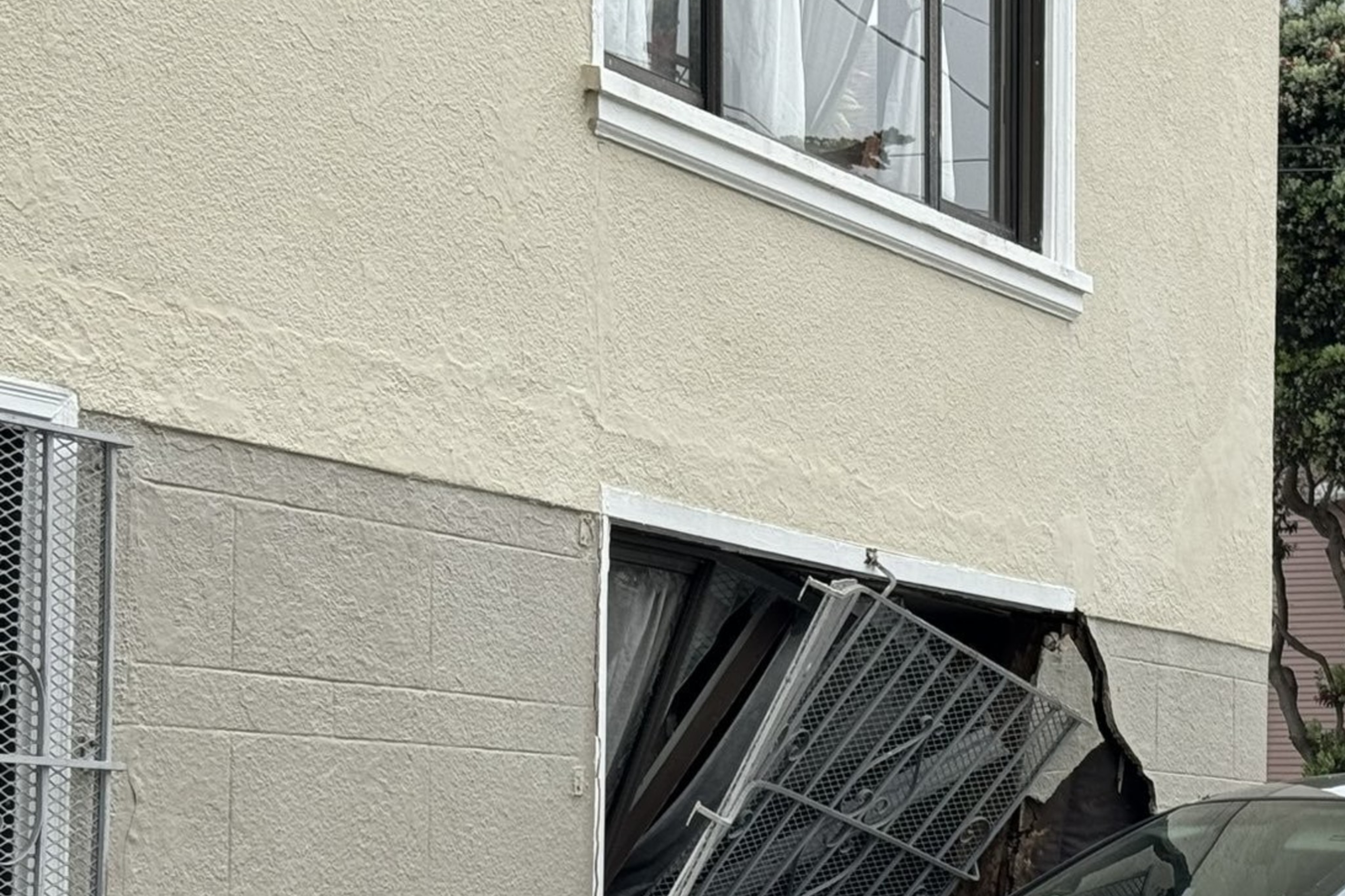 A gray car has crashed into the side of a building, damaging a garage door. A police officer is at the scene, inspecting the damage.