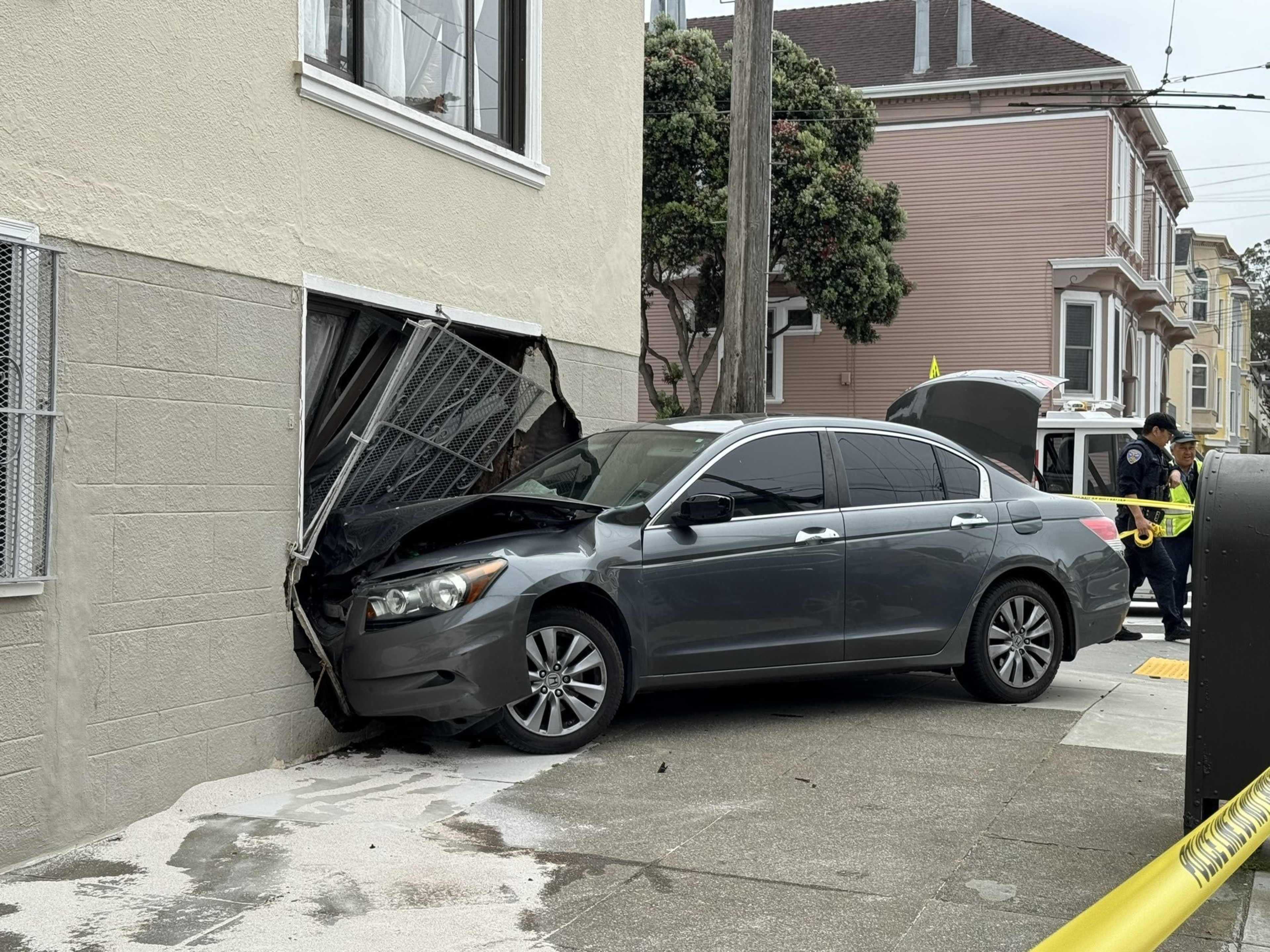 A gray car has crashed into the side of a building, damaging a garage door. A police officer is at the scene, inspecting the damage.