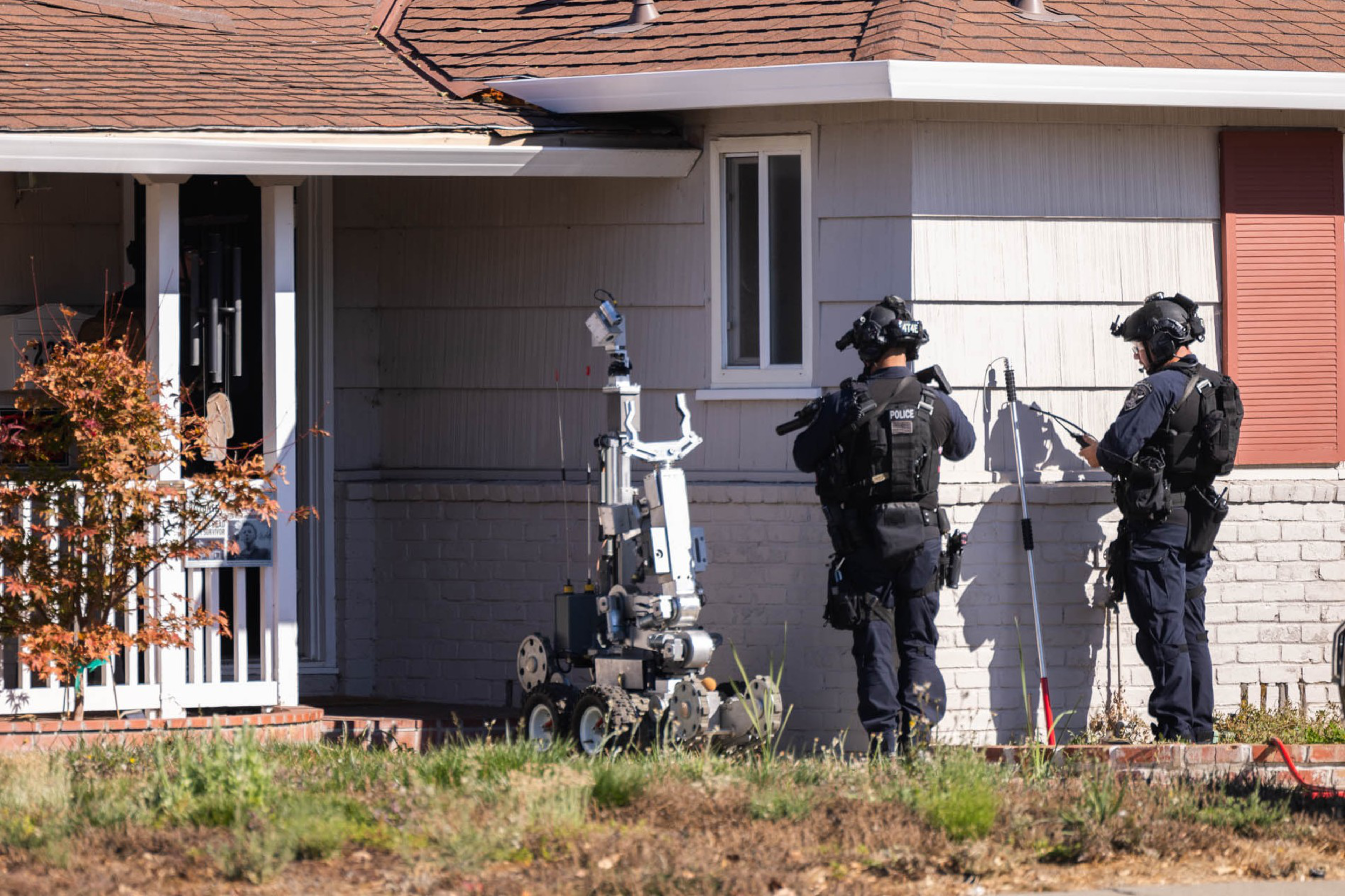 Two police officers in tactical gear operate a bomb disposal robot outside a suburban house with a brown roof and gray siding.