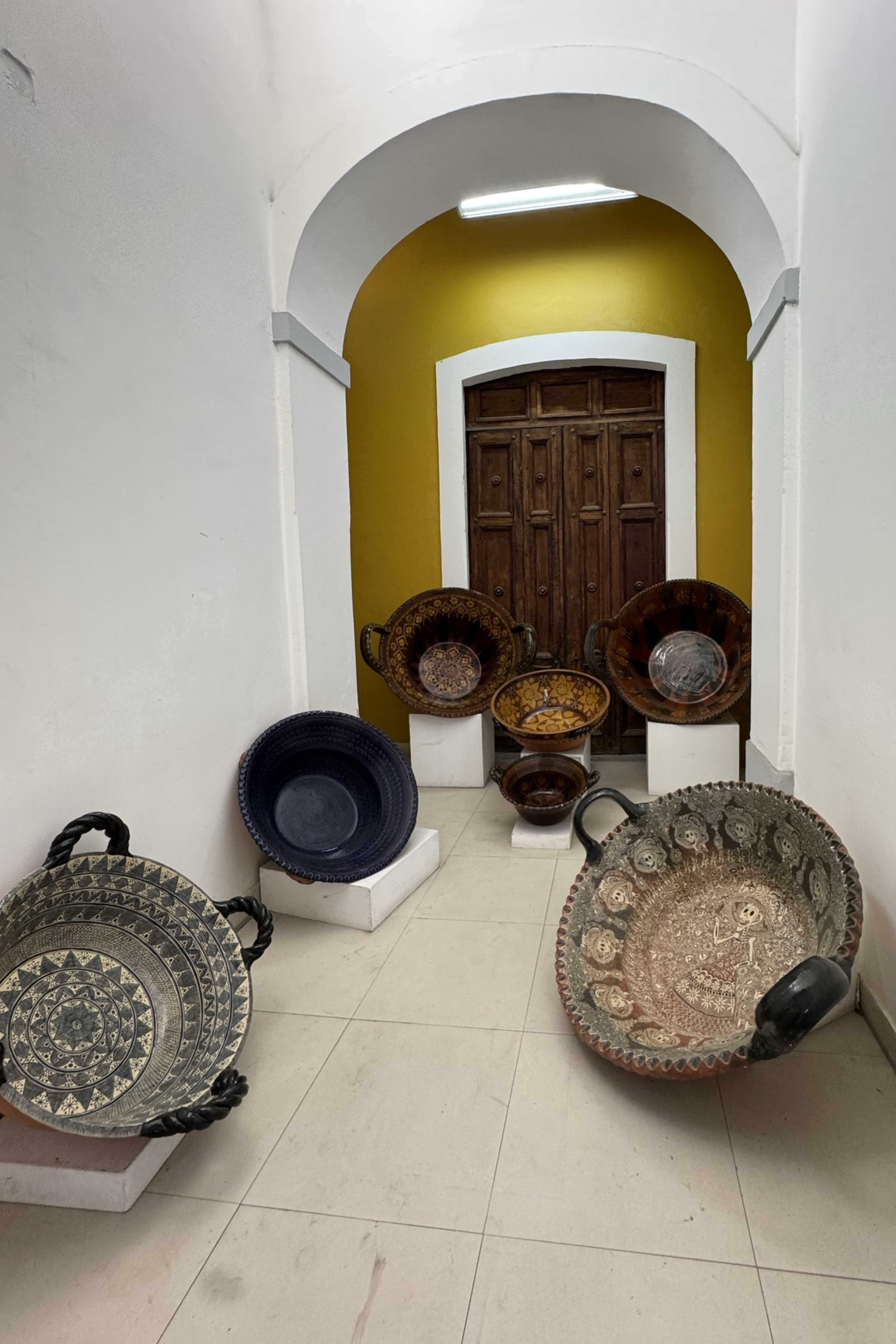 The image shows a narrow hallway with a yellow wall and wooden door at the end. Several large, intricately designed bowls with handles are displayed on the floor.