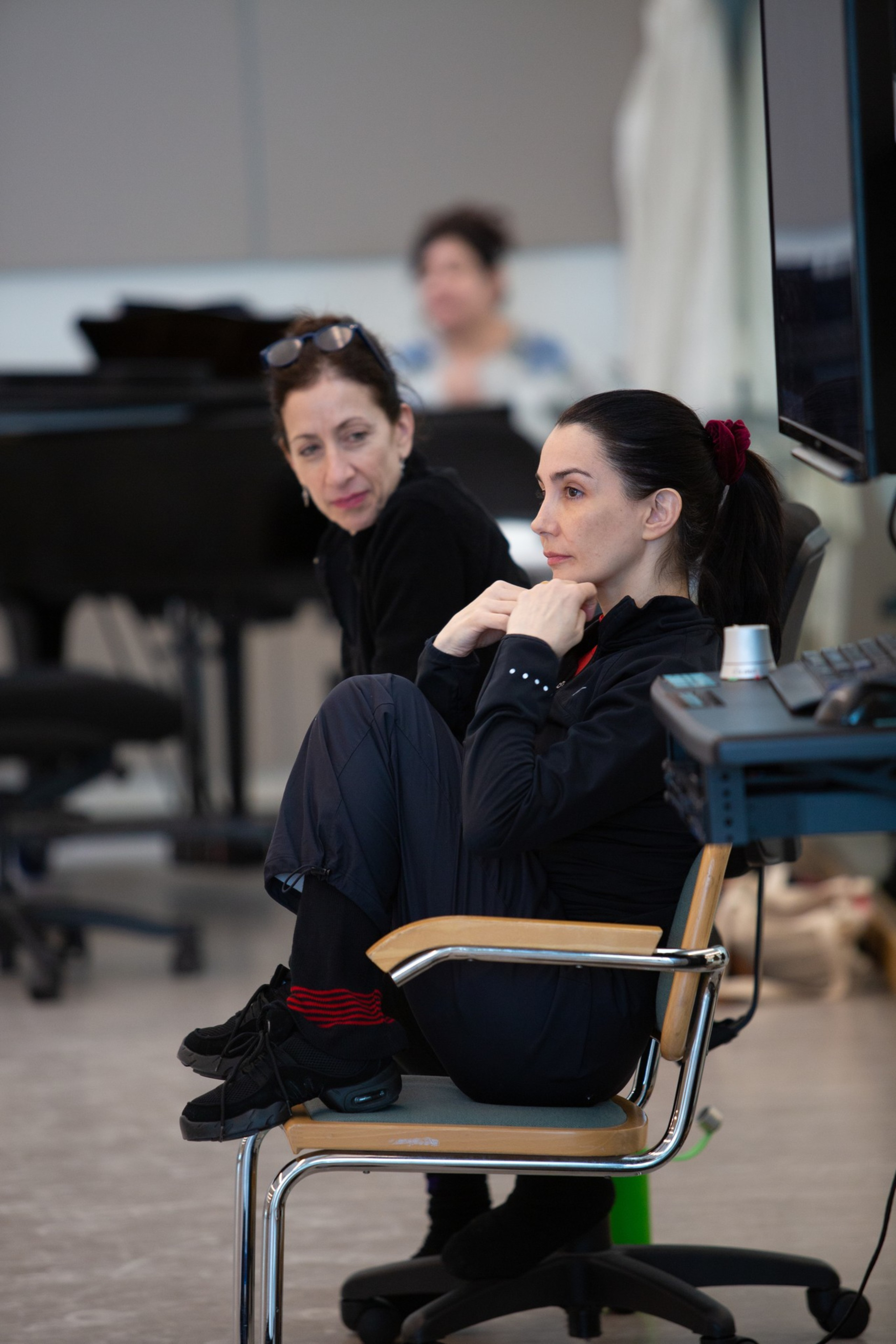 Two women sit near a piano; one is contemplative with a hand on her chin.