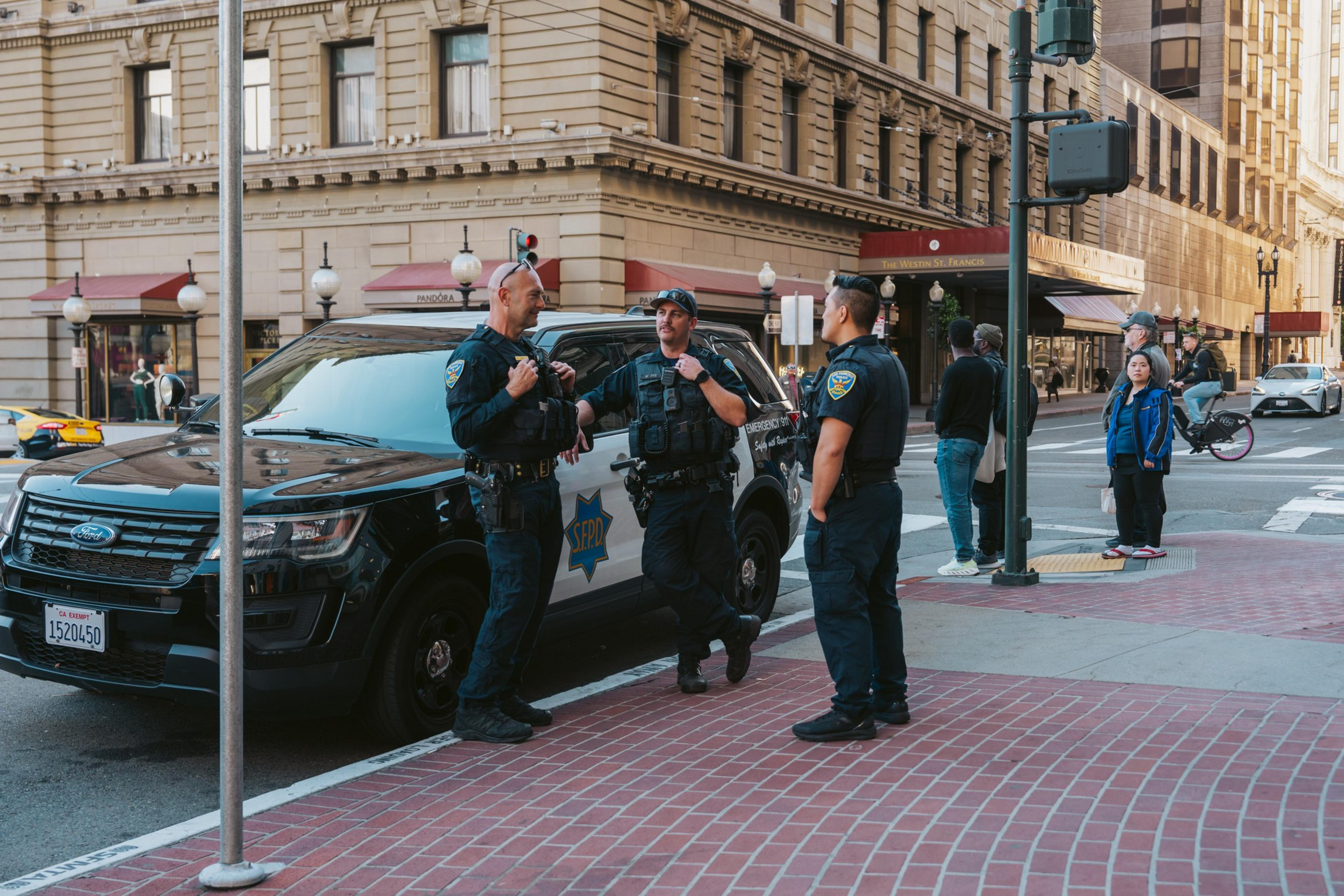 Three police officers stand talking beside a patrol car in a city street. Nearby, pedestrians, including a child, observe, and a cyclist passes by.