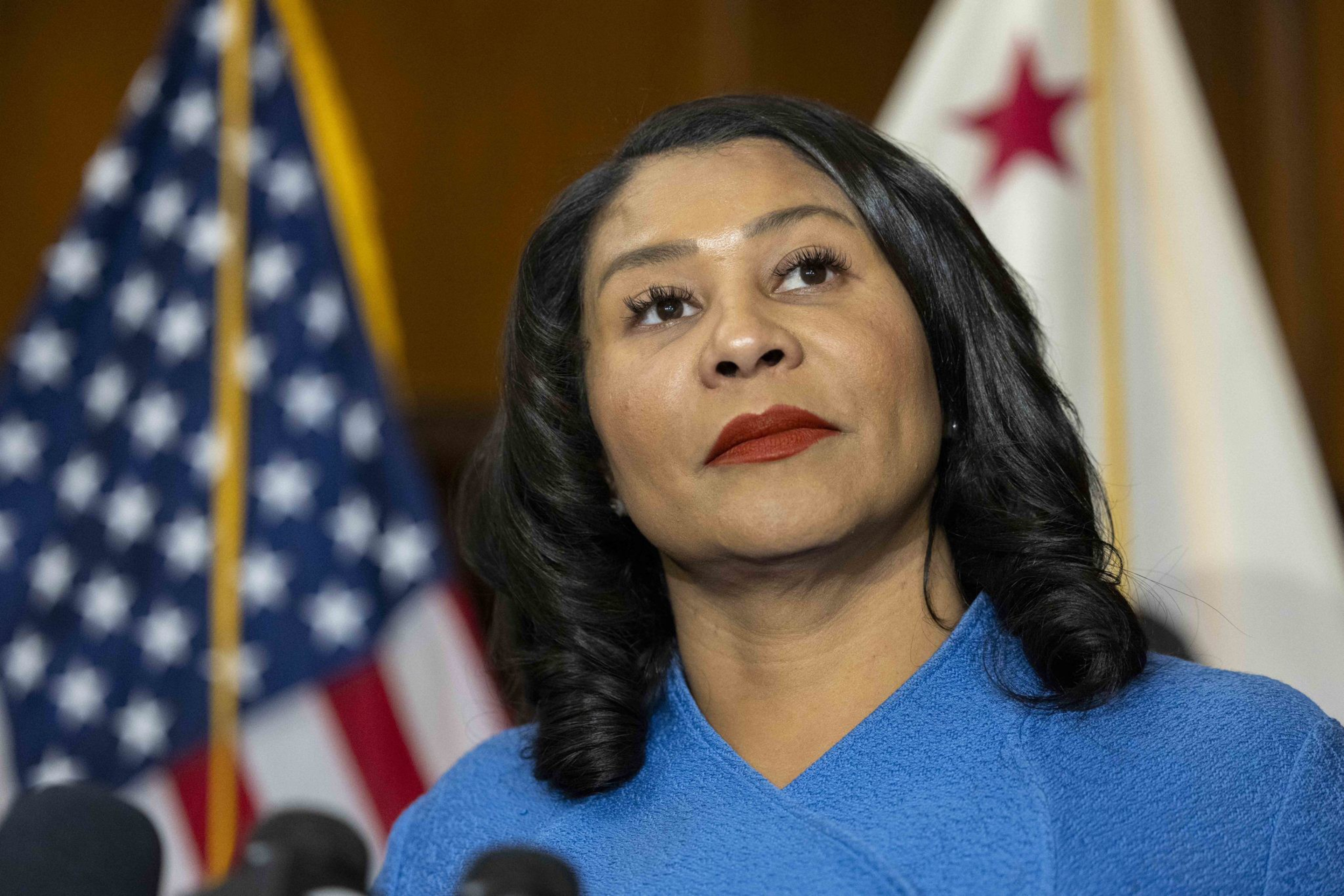A person in a blue outfit stands before microphones, with U.S. and California flags behind, appearing thoughtful and composed.