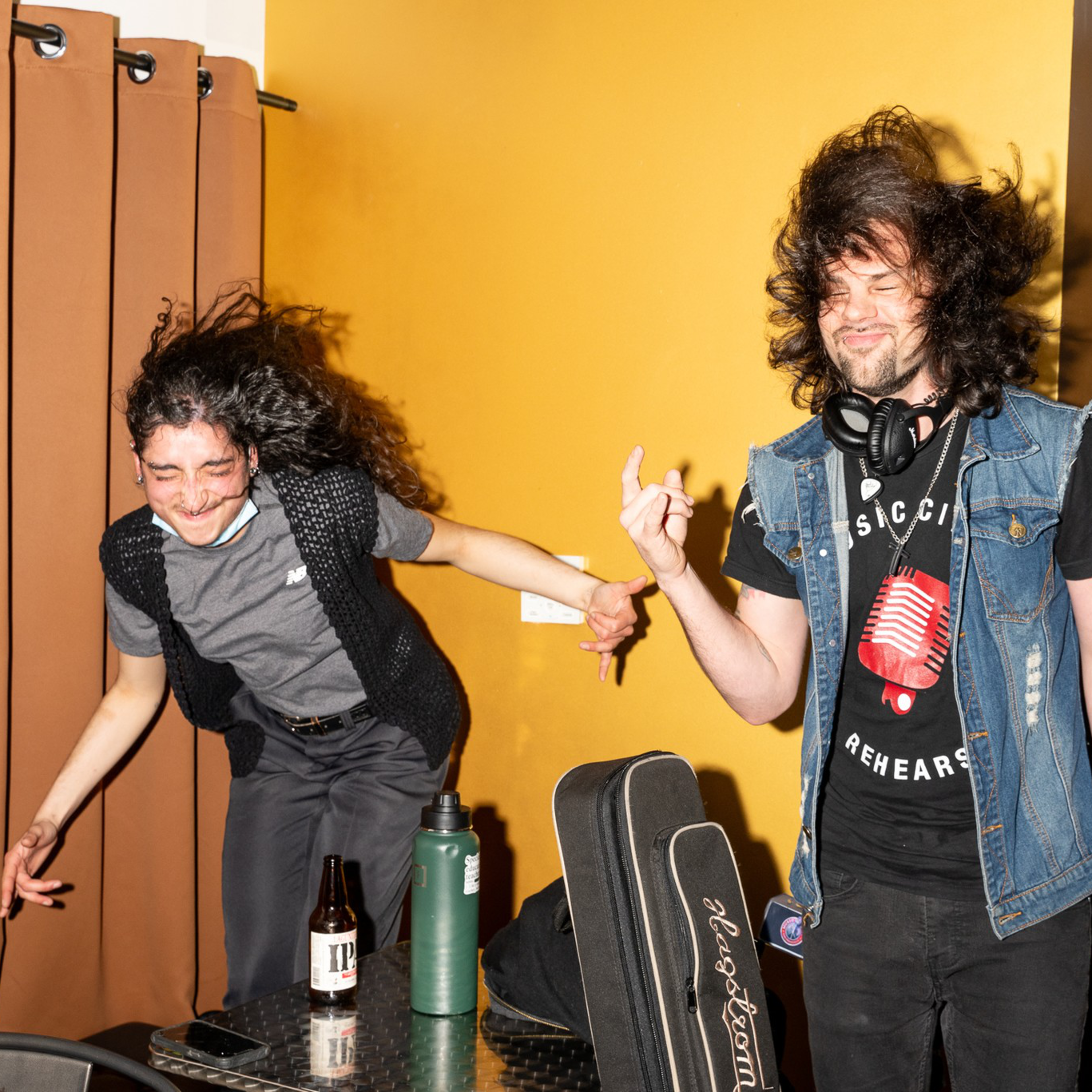 Two people are energetically dancing, hair flying, in front of a curtain. One holds a beer, the other a guitar case. A person sits in the background with a laptop.