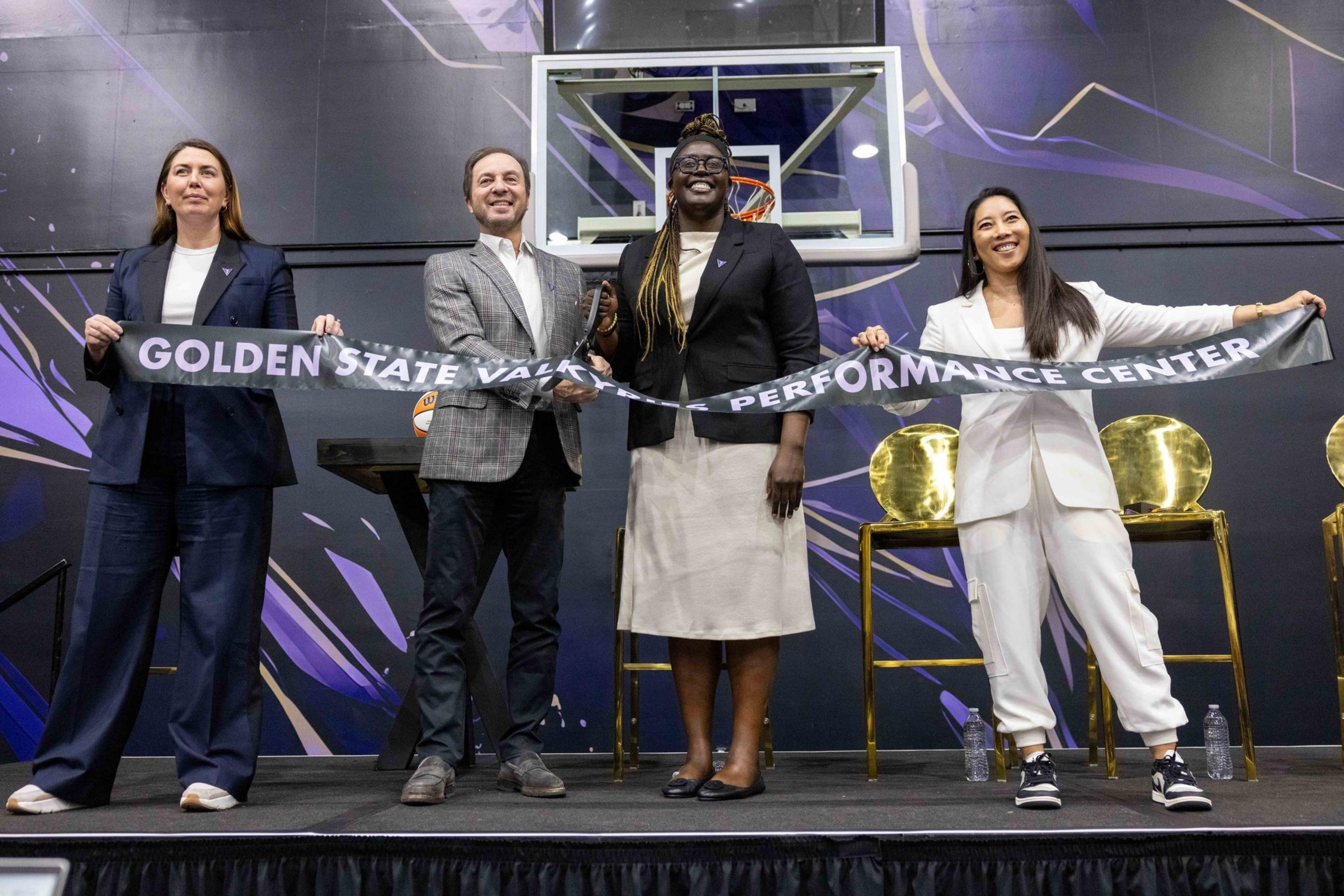 Four individuals hold a ribbon that reads &quot;Golden State Valkyries Performance Center&quot; in front of a basketball hoop, smiling, with three golden chairs behind them.