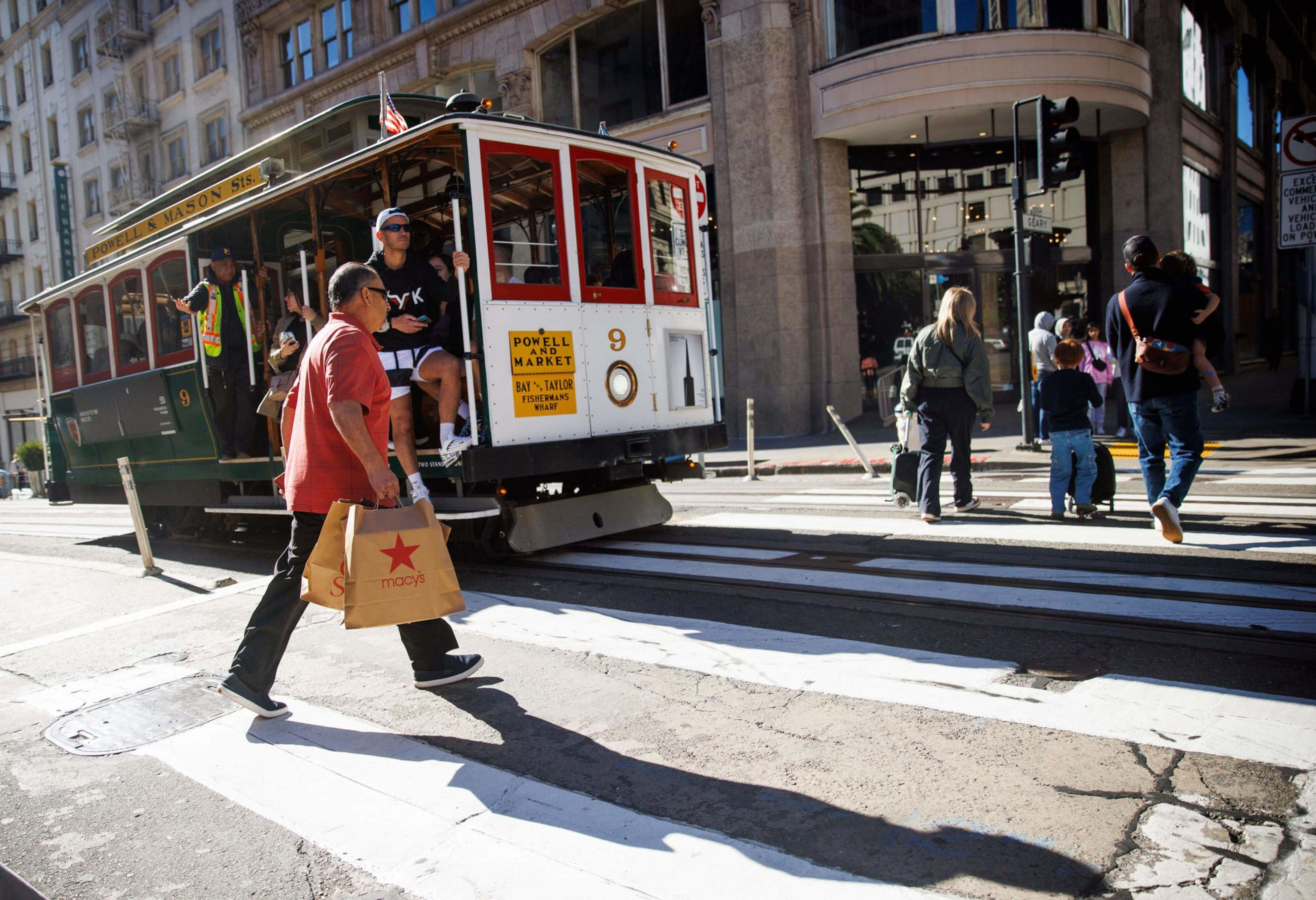 A man carrying Macy's shopping bags crosses a street in front of a historic San Francisco cable car, with other pedestrians walking nearby.