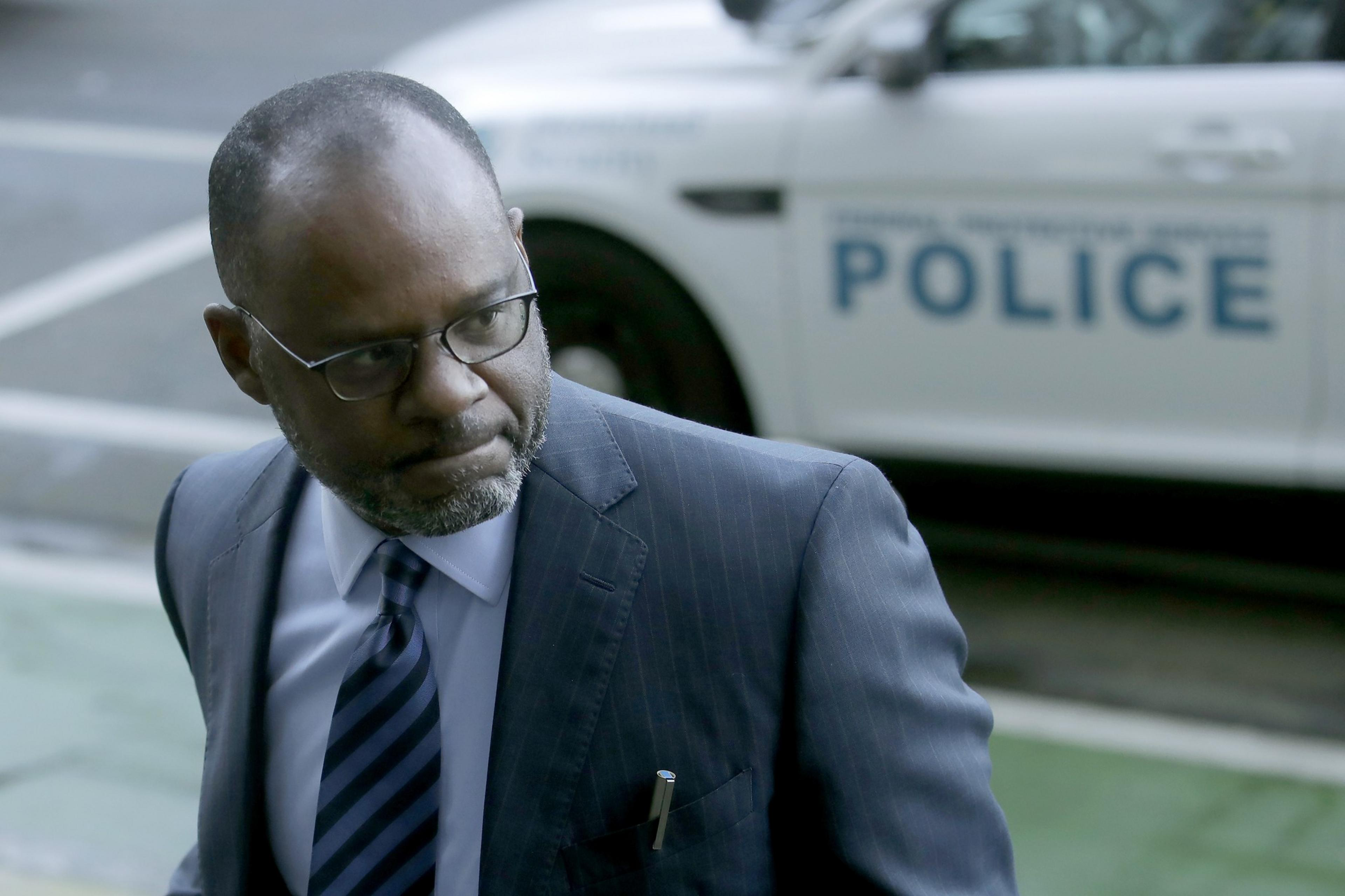 A serious man in glasses and a blue suit looks to his side while standing near a police car with the word "POLICE" visible in the background.