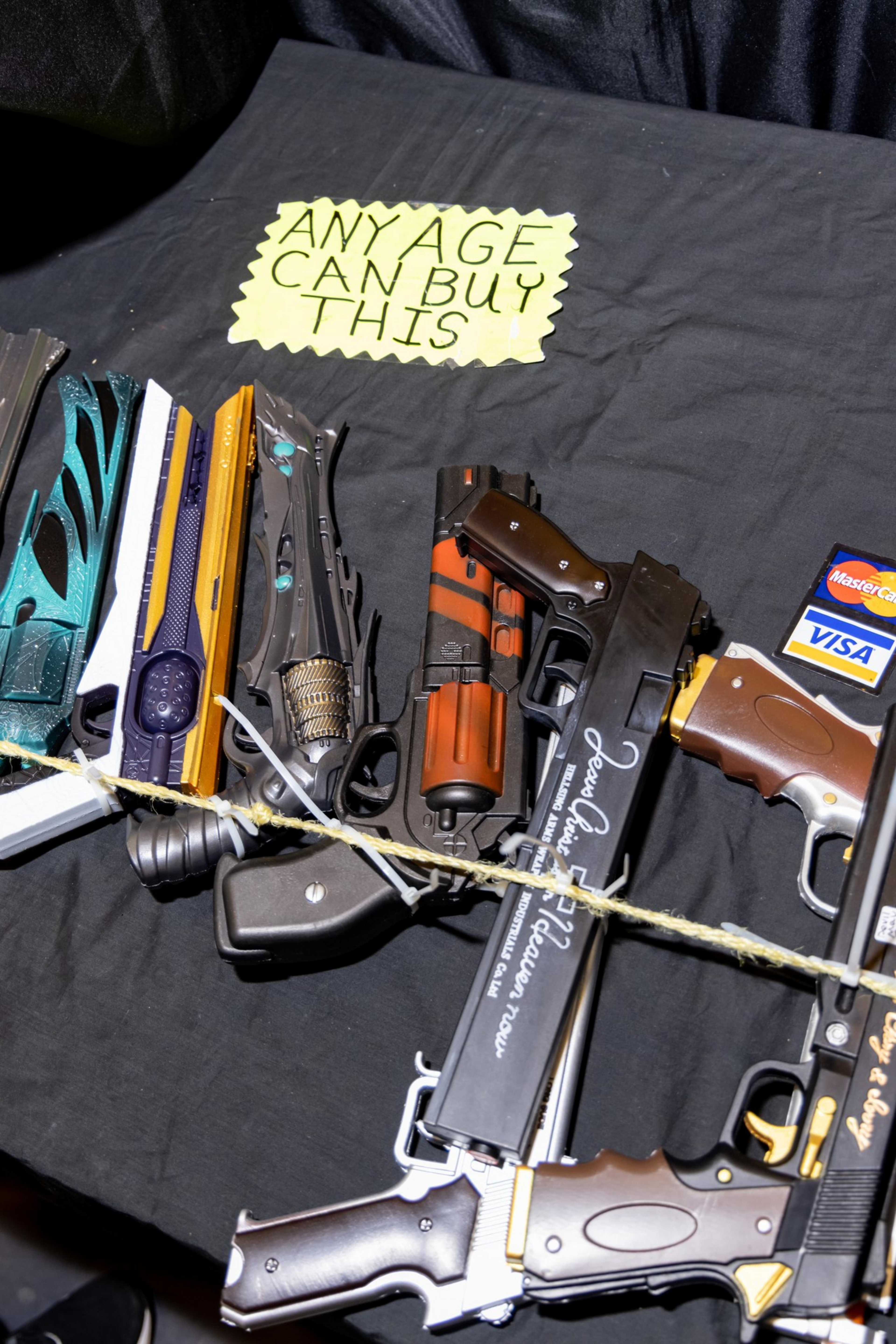 The image shows toy guns laid on a table with a handwritten sign saying &quot;ANY AGE CAN BUY THIS.&quot; There are also MasterCard and Visa card logos nearby.