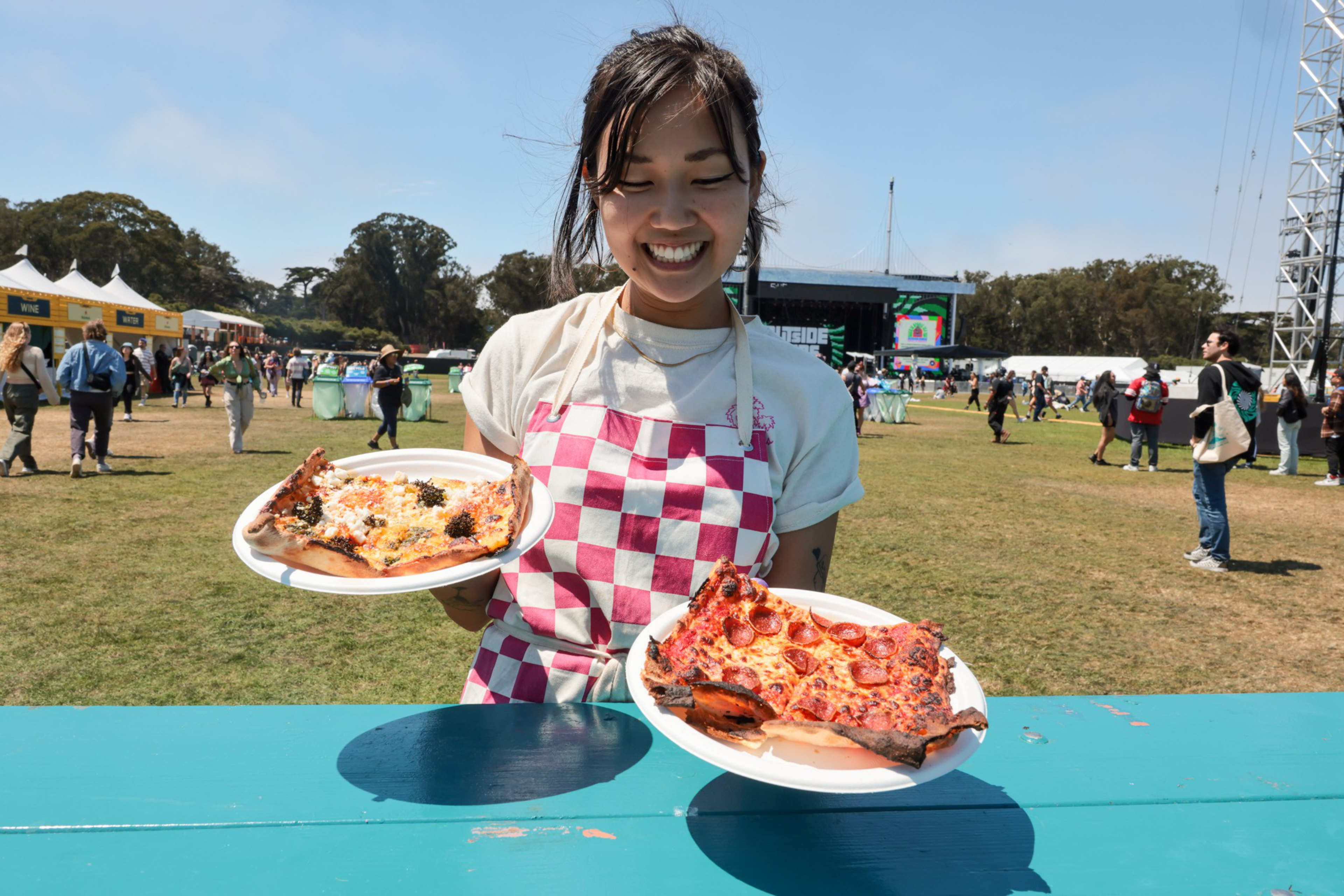 A smiling woman wearing a red and white checkered apron holds two plates with large pizza slices outdoors at a crowded event on a sunny day.