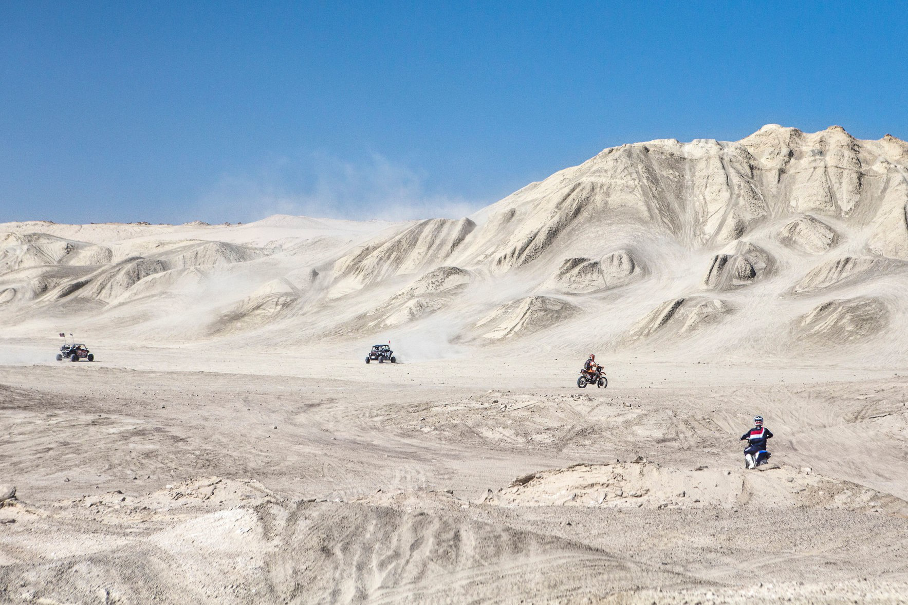Two dune buggies and two dirt bikers ride through a desert on a clear day.