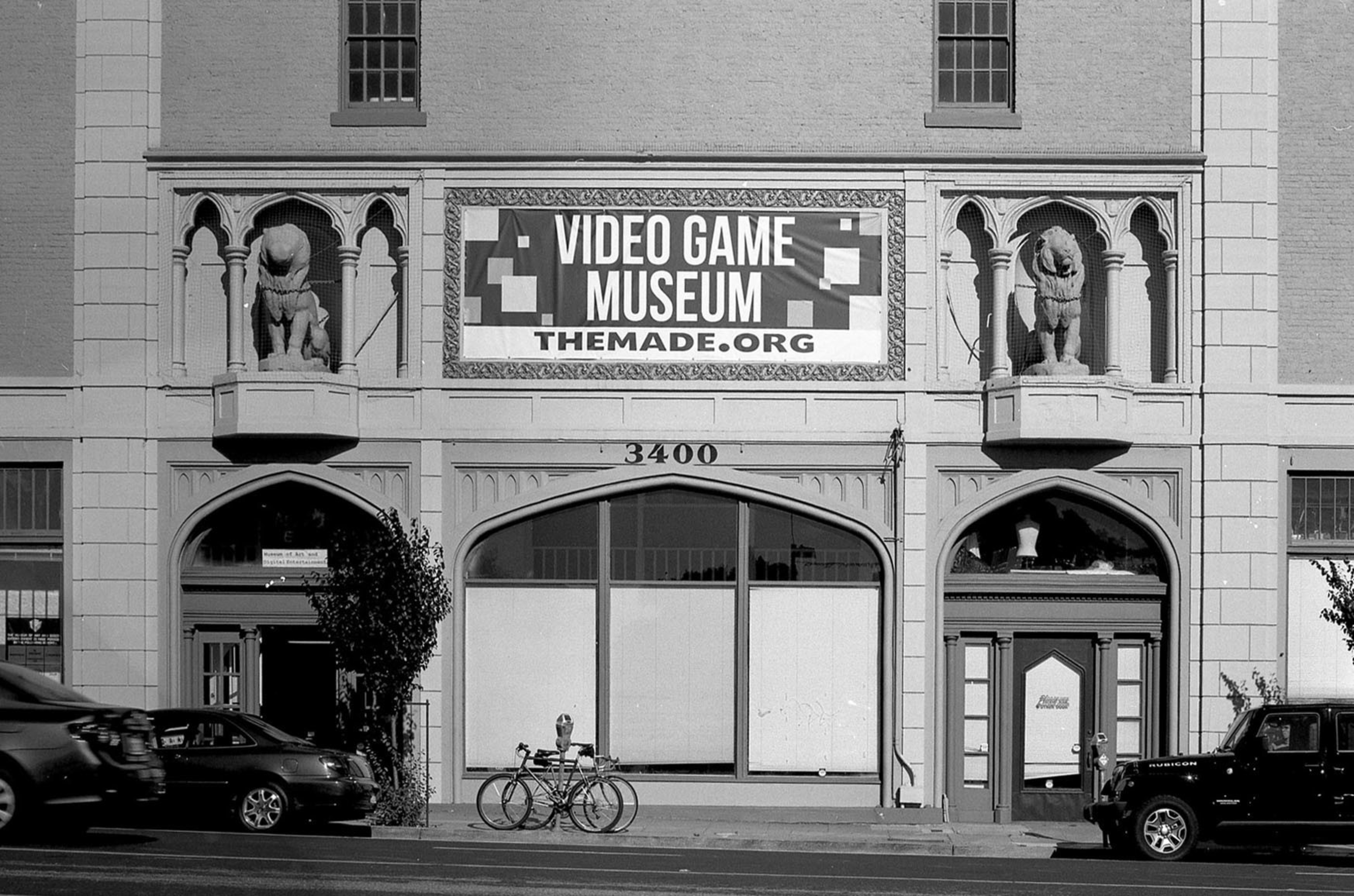A building with Gothic-style arches and lion statues displays a sign for the Video Game Museum at themade.org above the address number 3400.