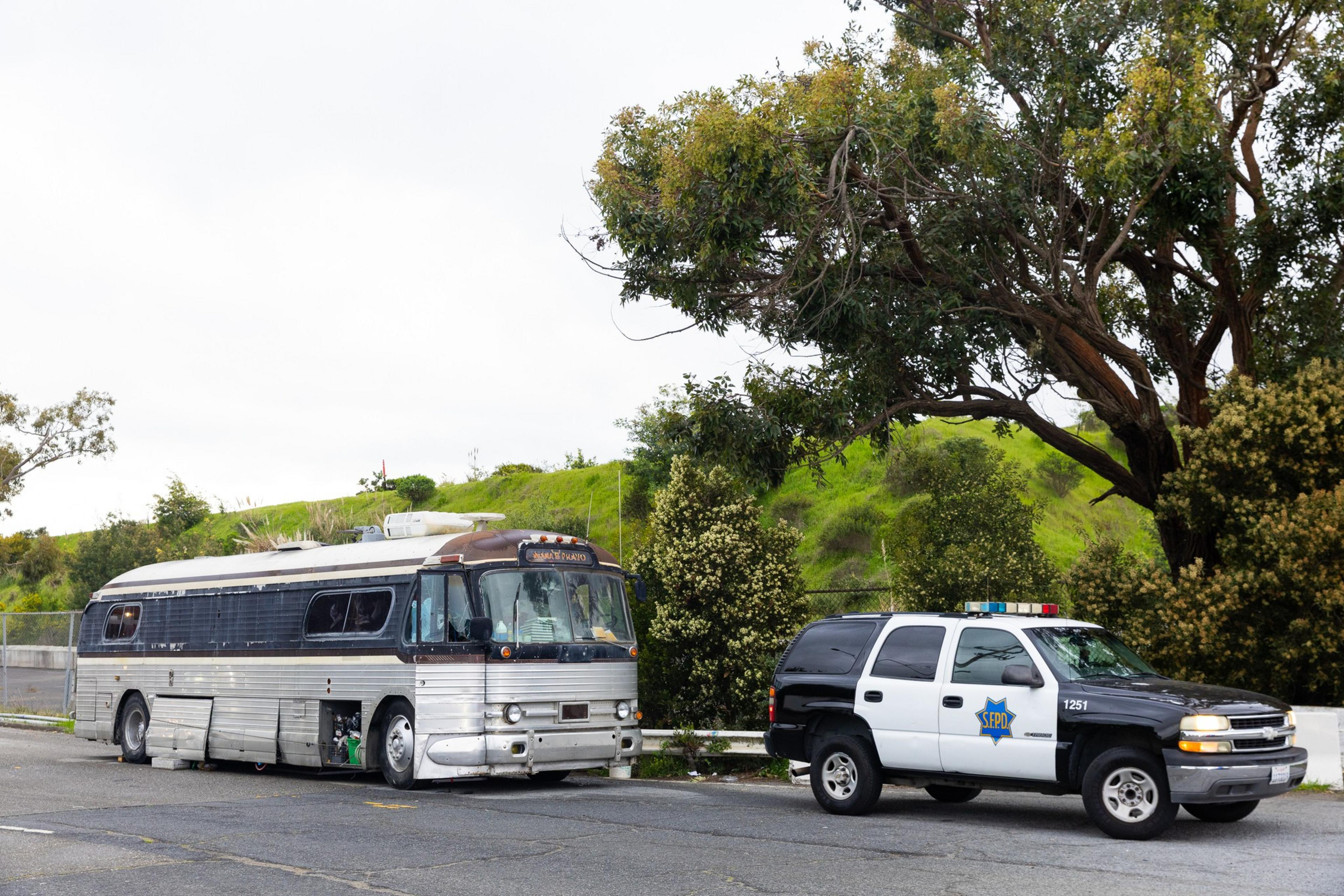A parked vintage-looking bus is beside a black-and-white police SUV with SFPD markings on a street lined with green hills and trees.