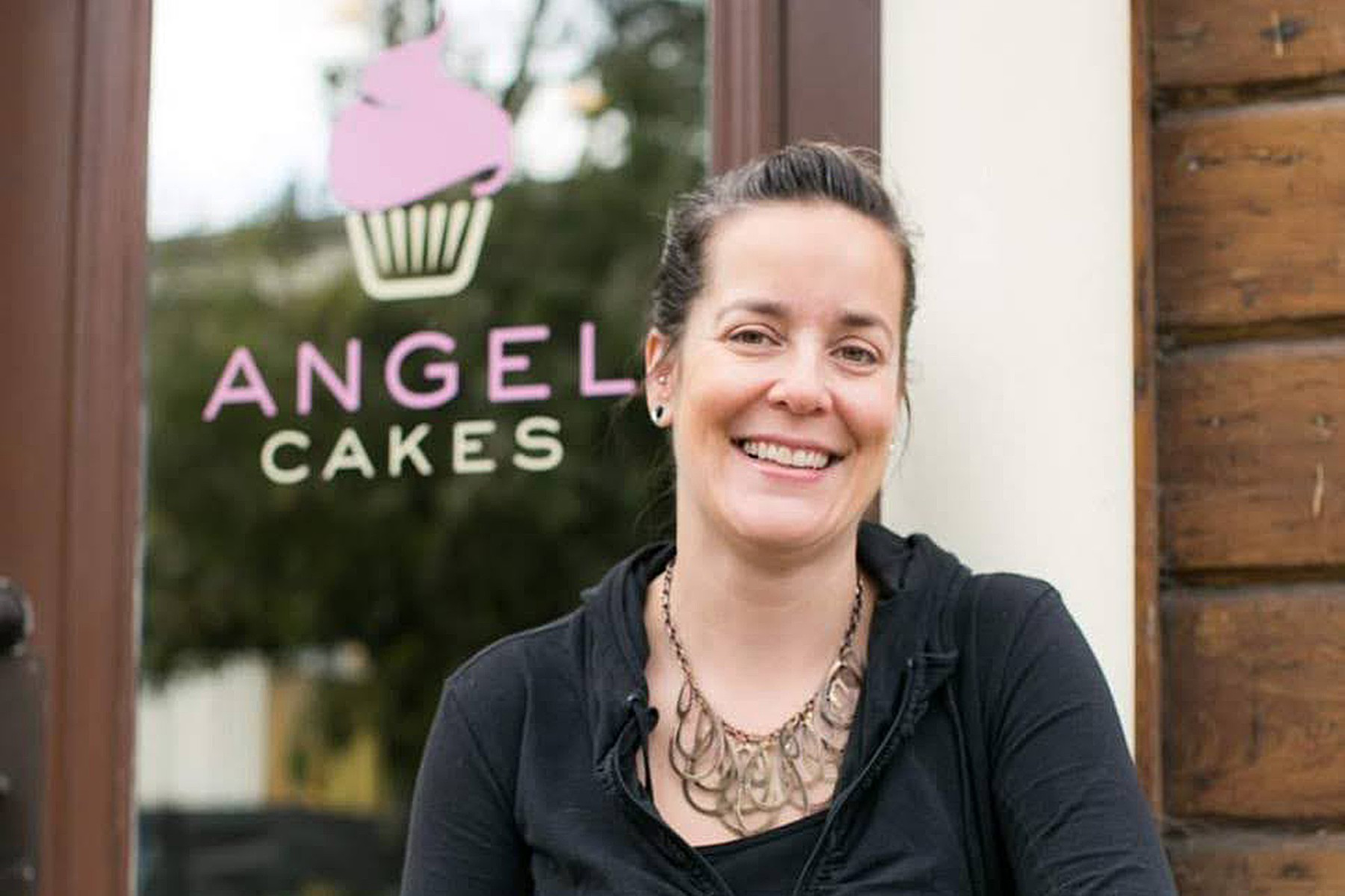 A smiling woman with dark hair pulled back, wearing a black top and necklace, stands in front of a glass door with a pink cupcake logo and "Angel Cakes" sign.