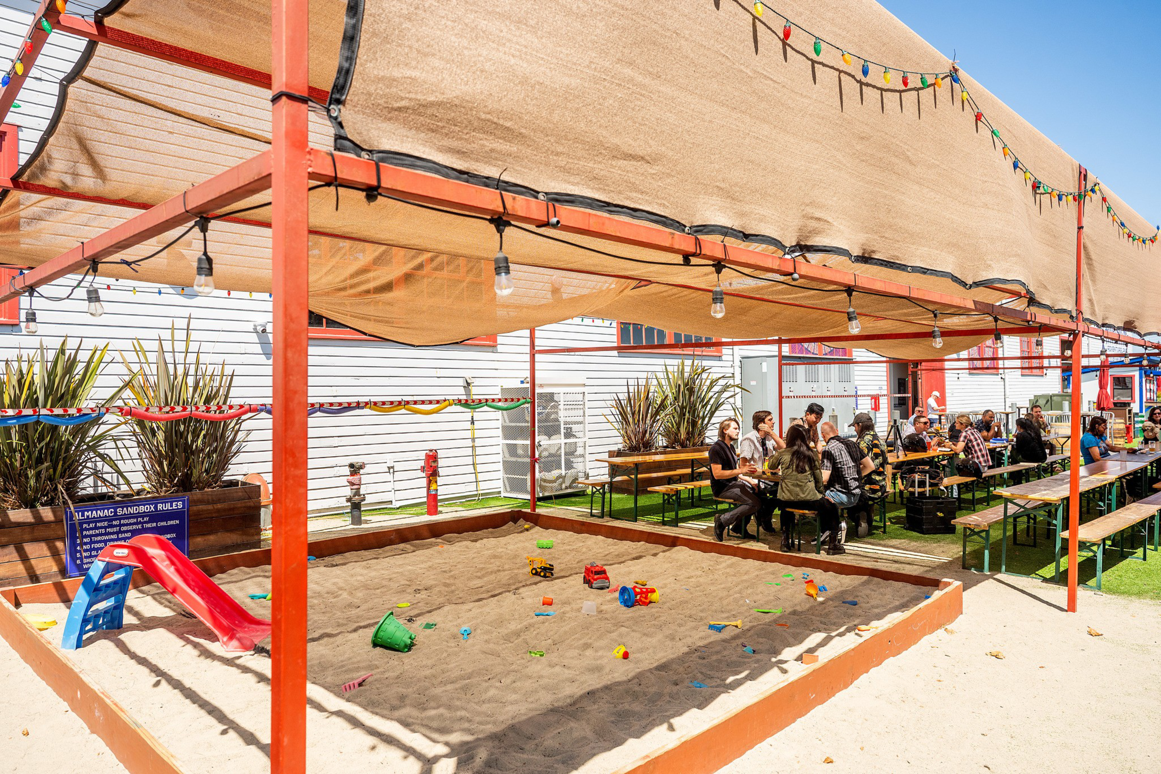 A covered sandbox with scattered toys sits next to picnic tables where people are seated and chatting outdoors under string lights.