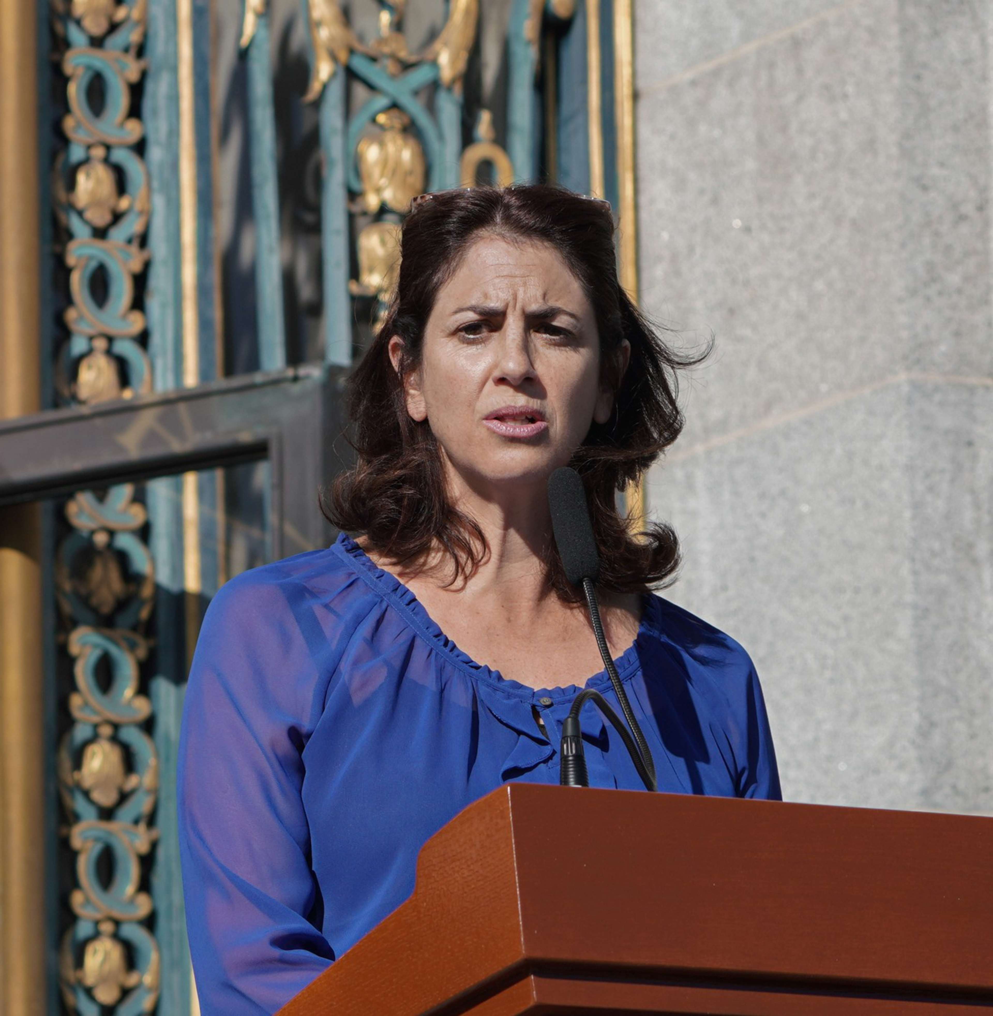 A woman with dark hair, wearing a blue top, is speaking at a podium with a microphone in front of a decorative building facade.