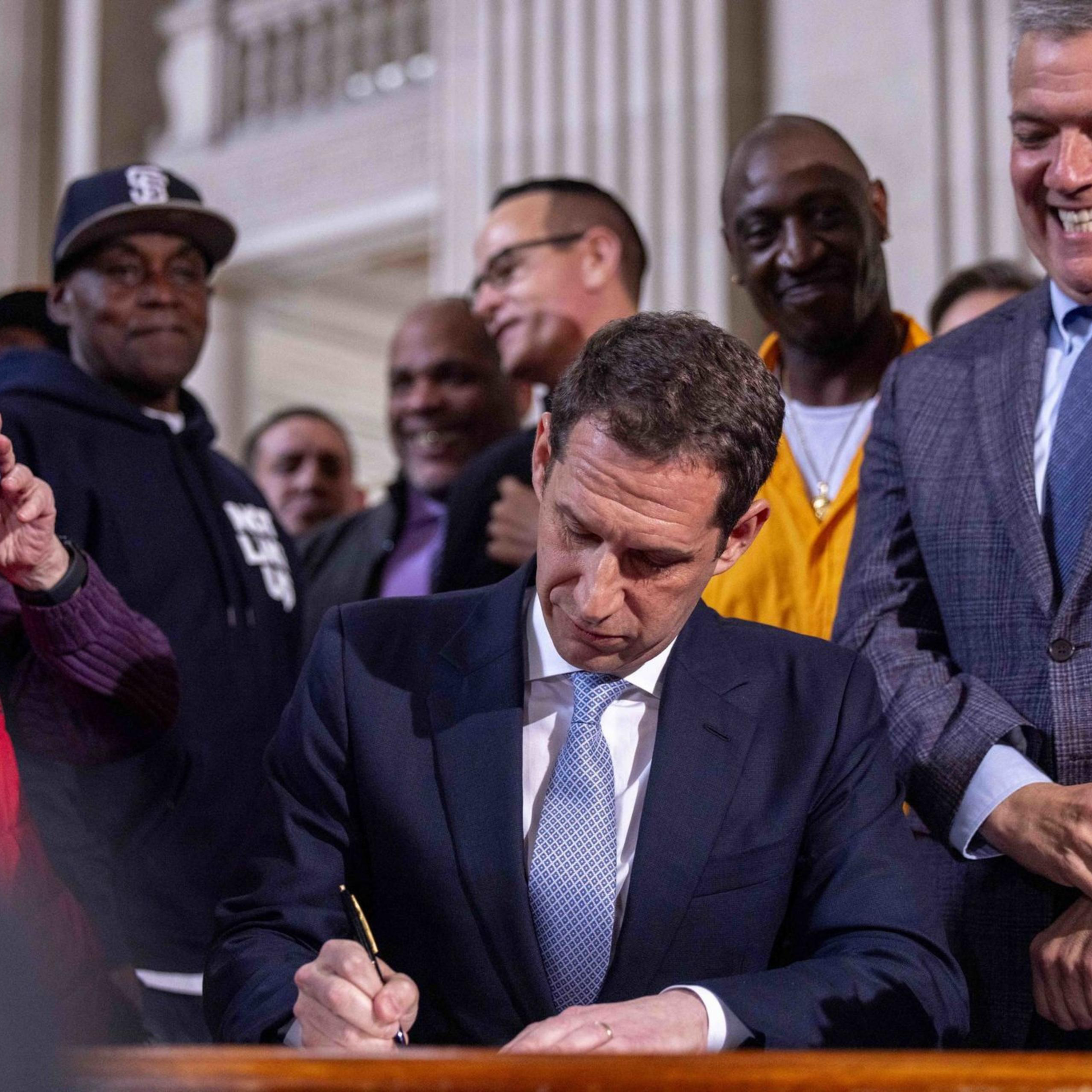 A man in a suit is signing a document at a desk, surrounded by a smiling group of people, including someone taking a photo with a smartphone.
