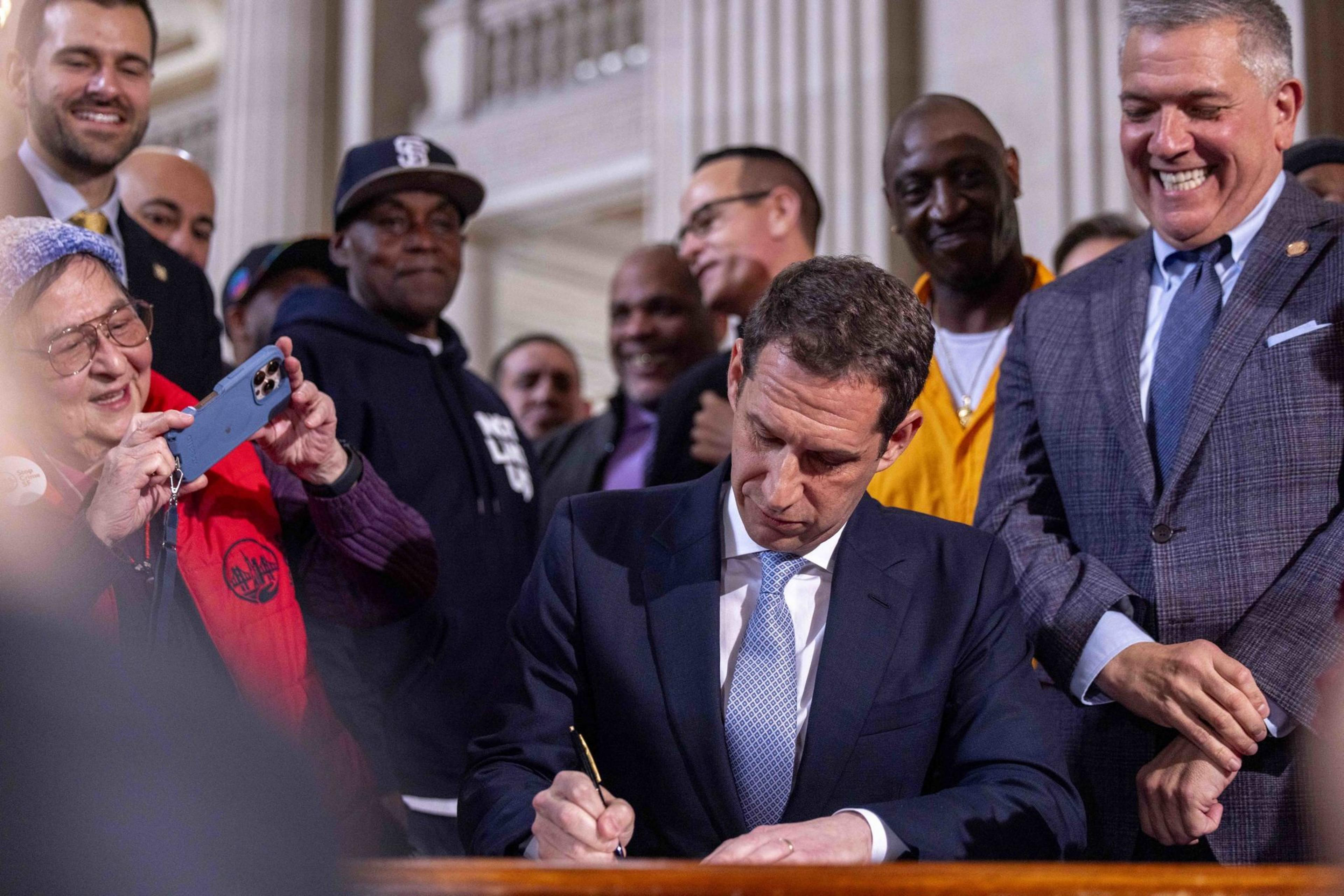 A man in a suit is signing a document at a desk, surrounded by a smiling group of people, including someone taking a photo with a smartphone.