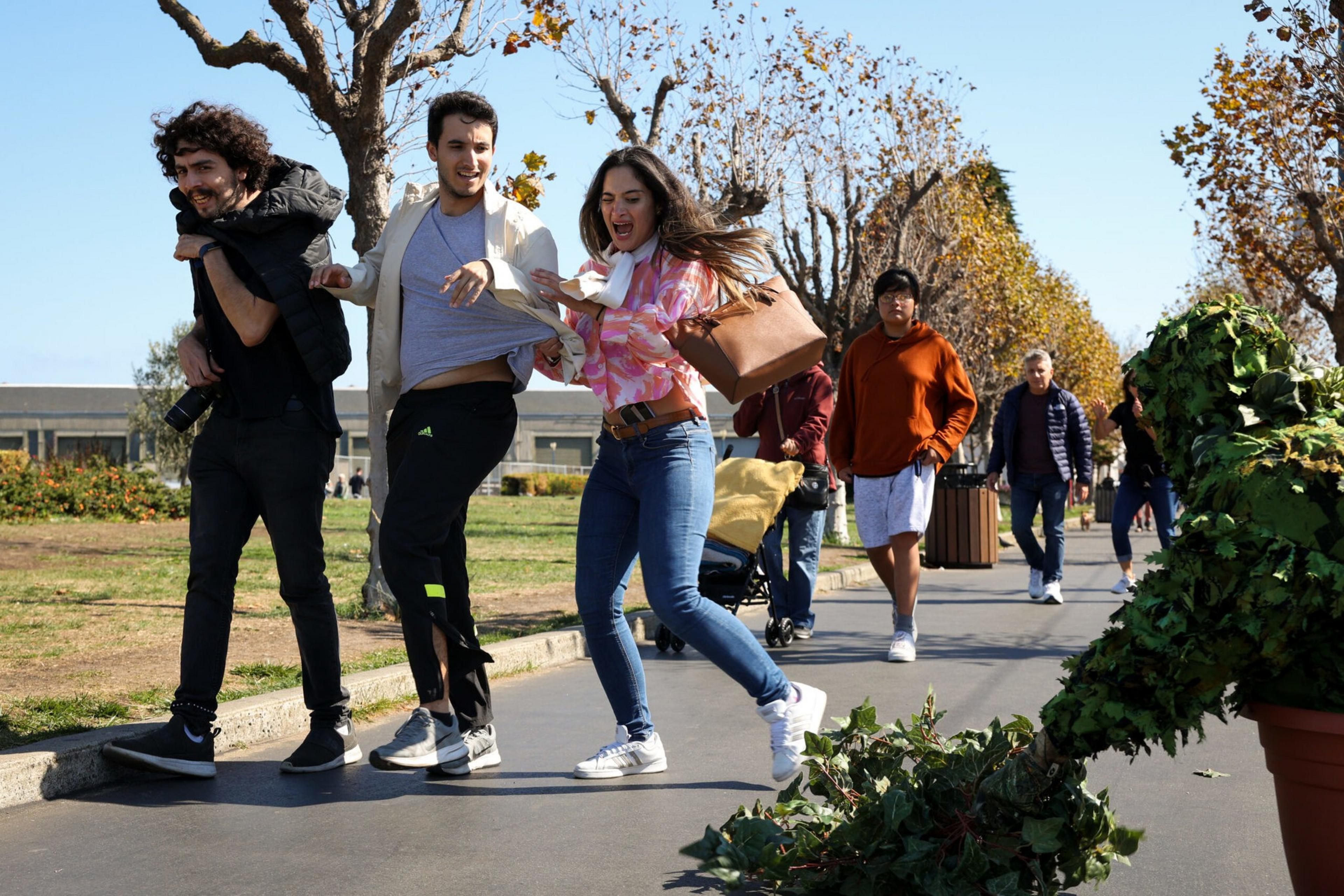 Three people react with surprise and laughter as a person in a leafy plant costume approaches them on a sunny park pathway.