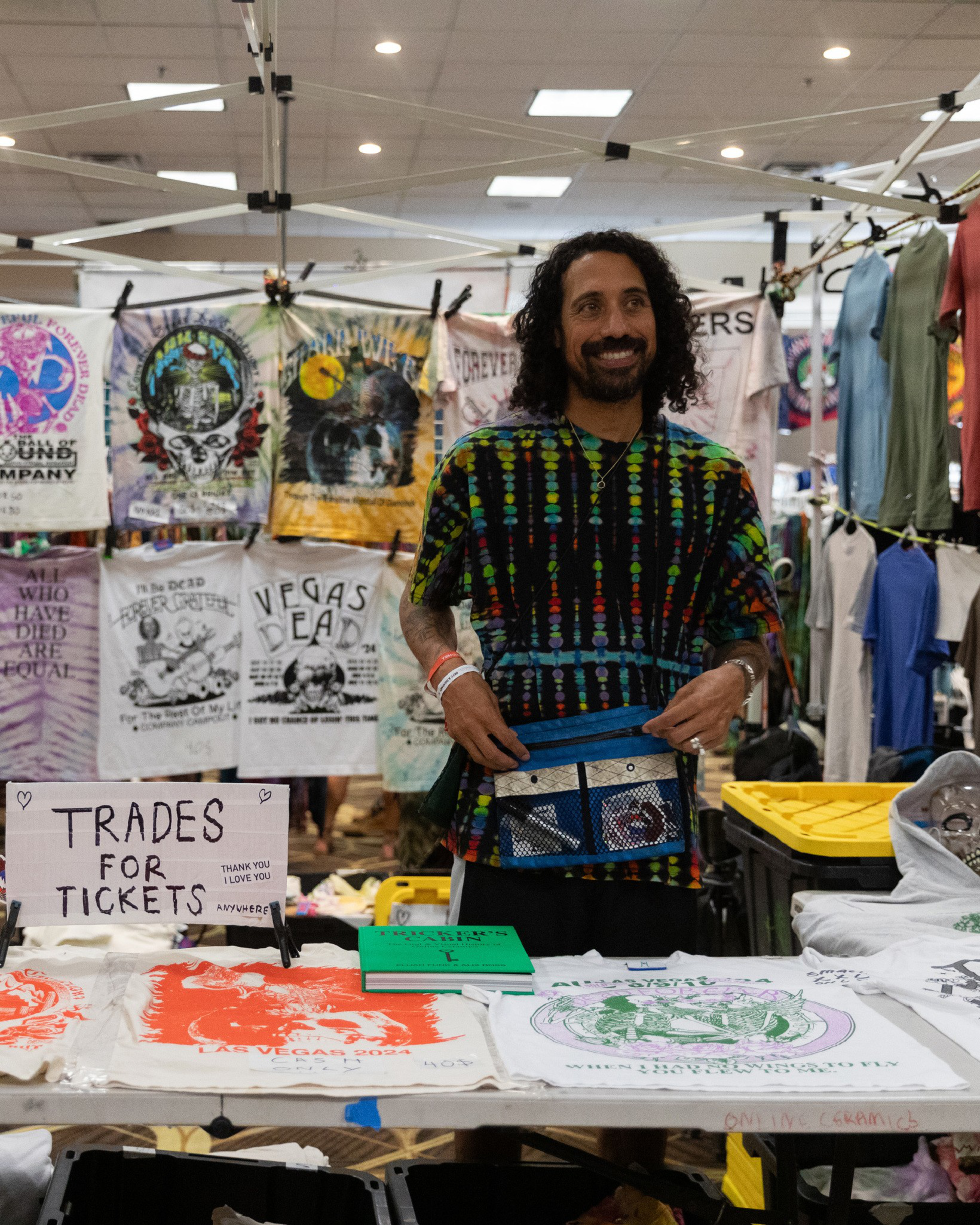A smiling man stands behind a table with T-shirts and a sign reading &quot;Trades for Tickets.&quot; He is in a booth displaying various colorful, printed T-shirts and garments.