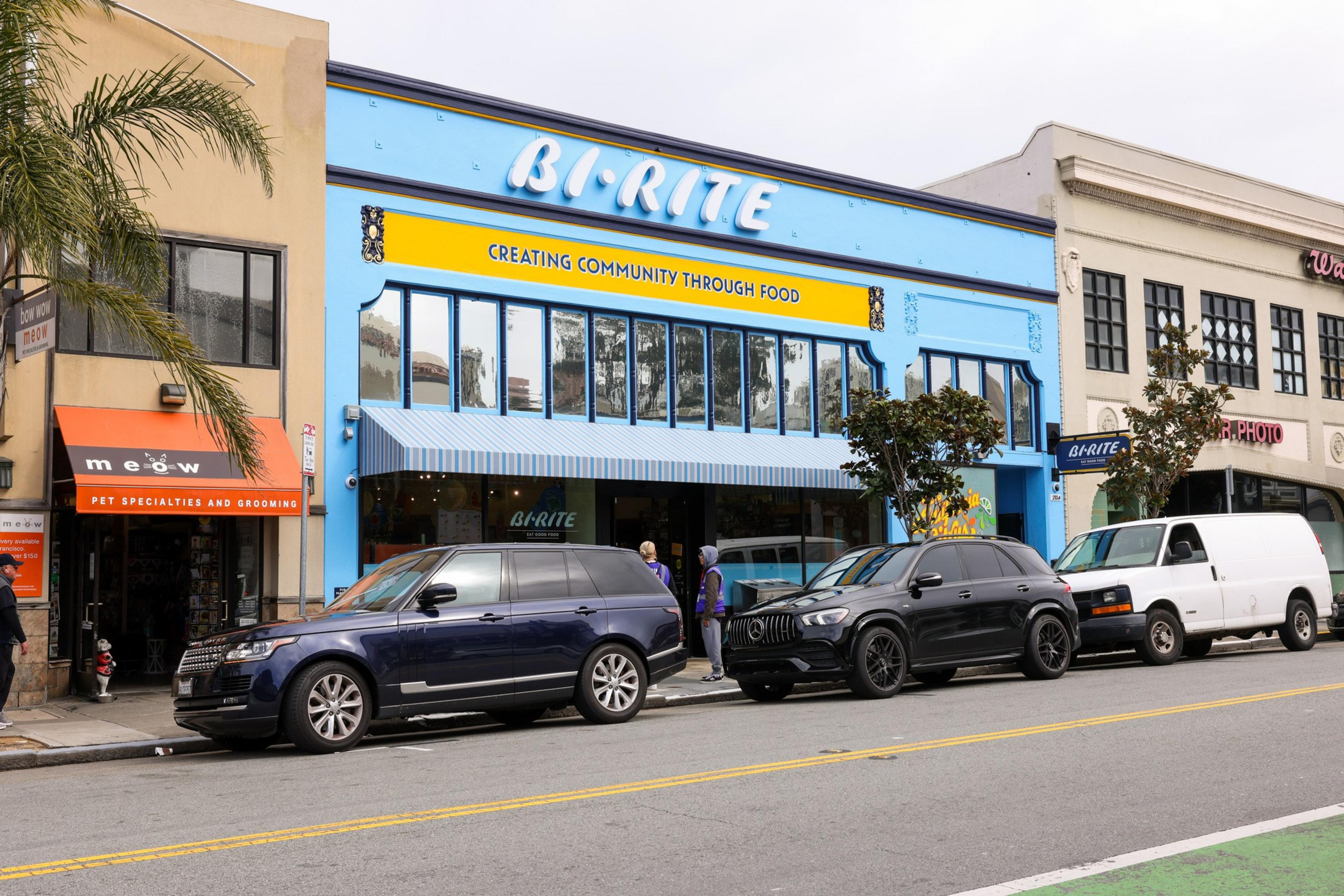 A vibrant blue and yellow building with &quot;Bi-Rite&quot; and &quot;Creating Community Through Food&quot; signage. Cars are parked in front, and a few people are nearby.