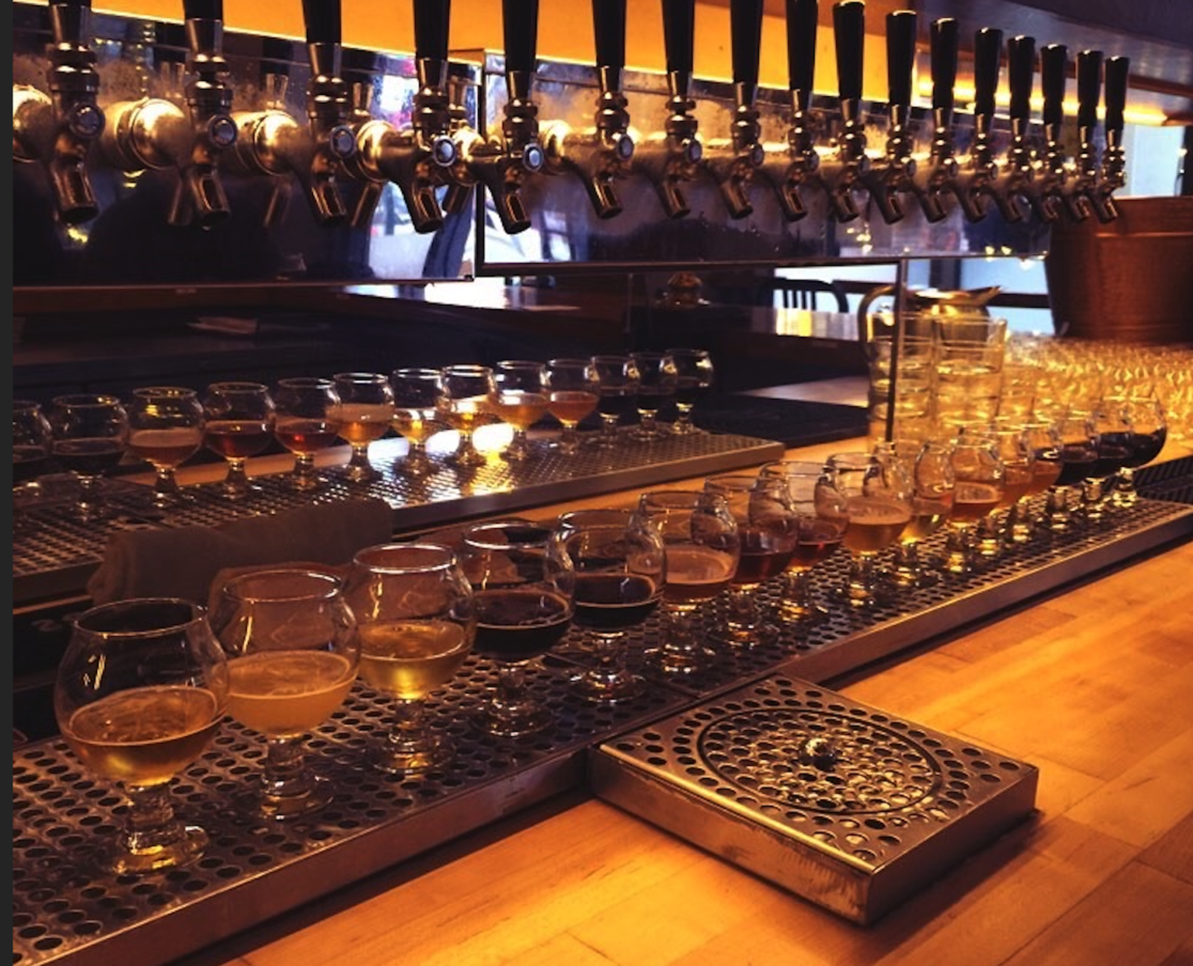 Twenty glasses of beer, each with different colors and foam levels, are lined up on a bar counter beneath numerous beer taps.