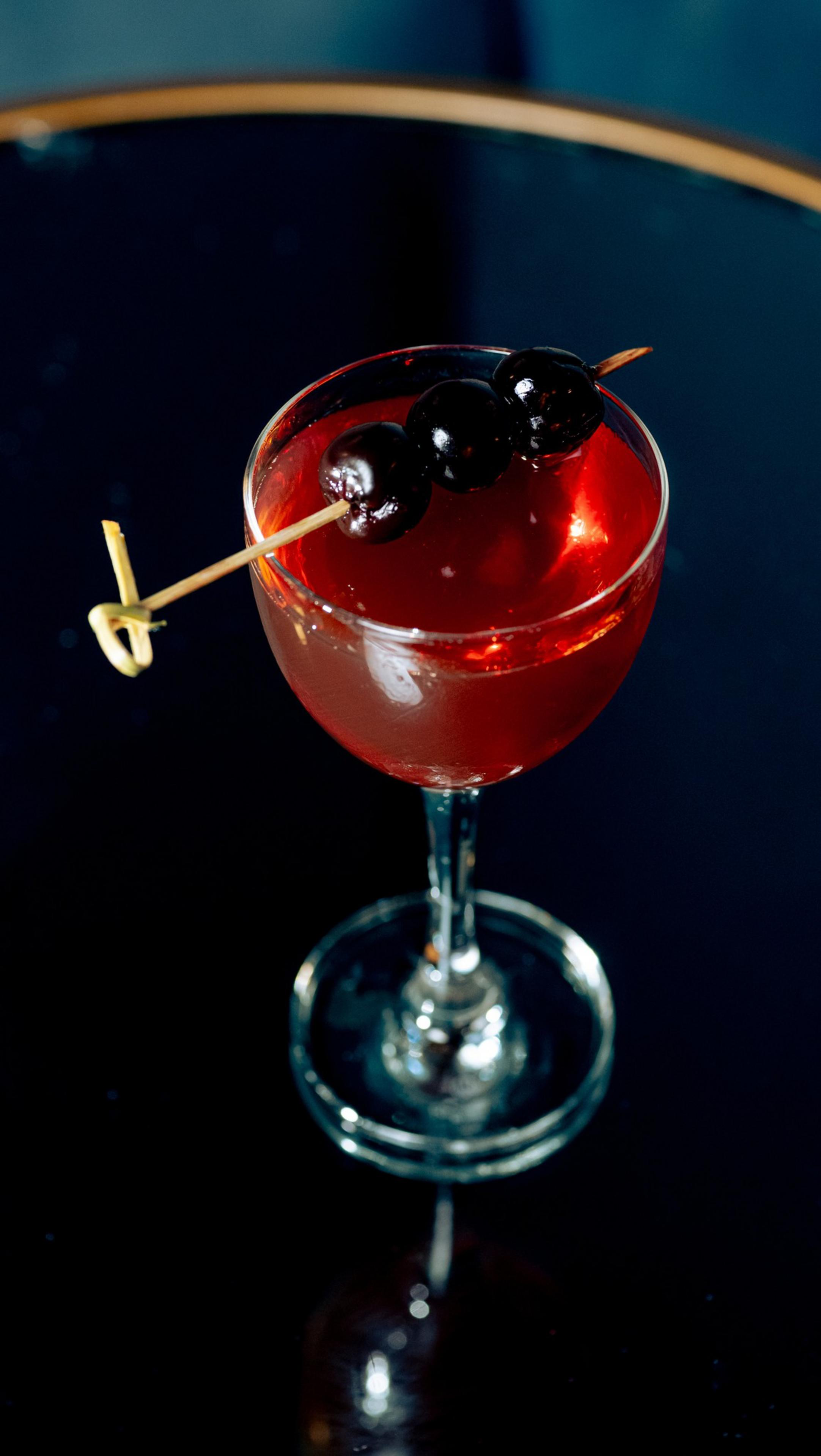 A red cocktail in a clear stemmed glass is garnished with three dark cherries on a wooden pick, placed on a reflective dark surface.