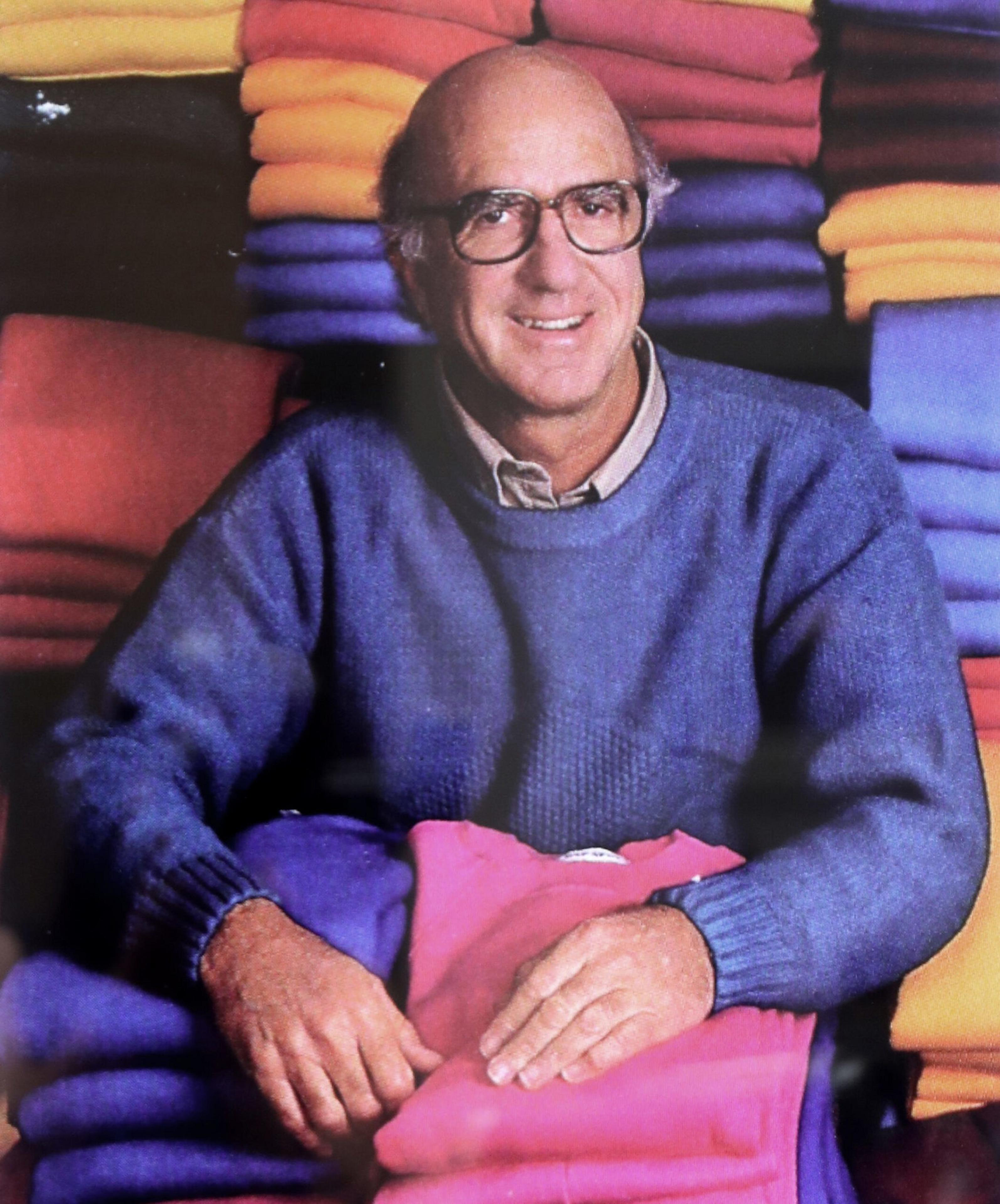 A smiling man wearing glasses and a blue sweater sits among neatly folded colorful sweaters stacked behind and beneath him.
