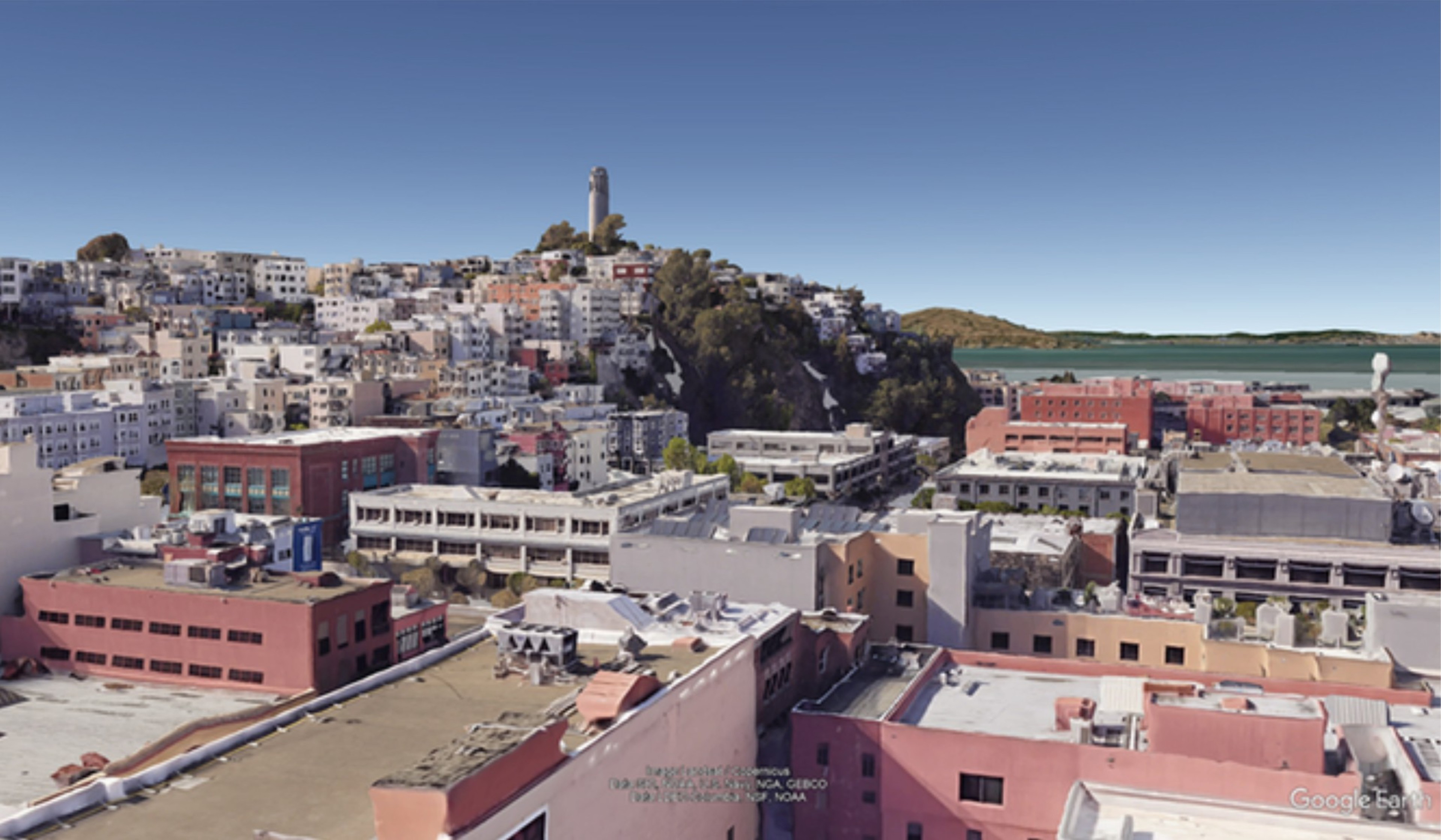 The image shows a cityscape with densely packed buildings climbing up a hill, topped by a tall tower. Below, more buildings are visible with a body of water in the background.