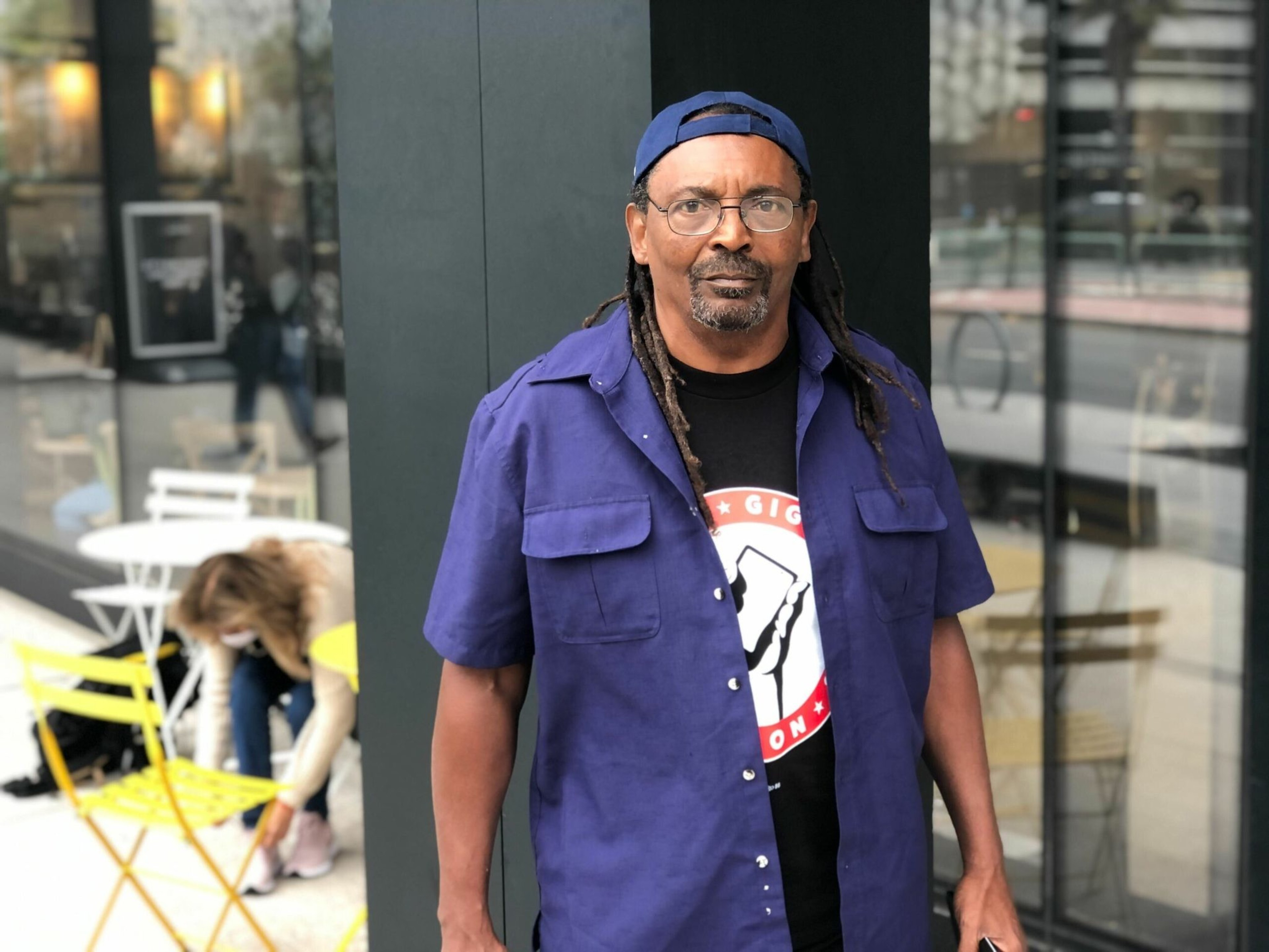 A man with glasses and dreadlocks wearing a blue cap backward, a blue short-sleeve shirt, and a black t-shirt stands outside near a glass wall.
