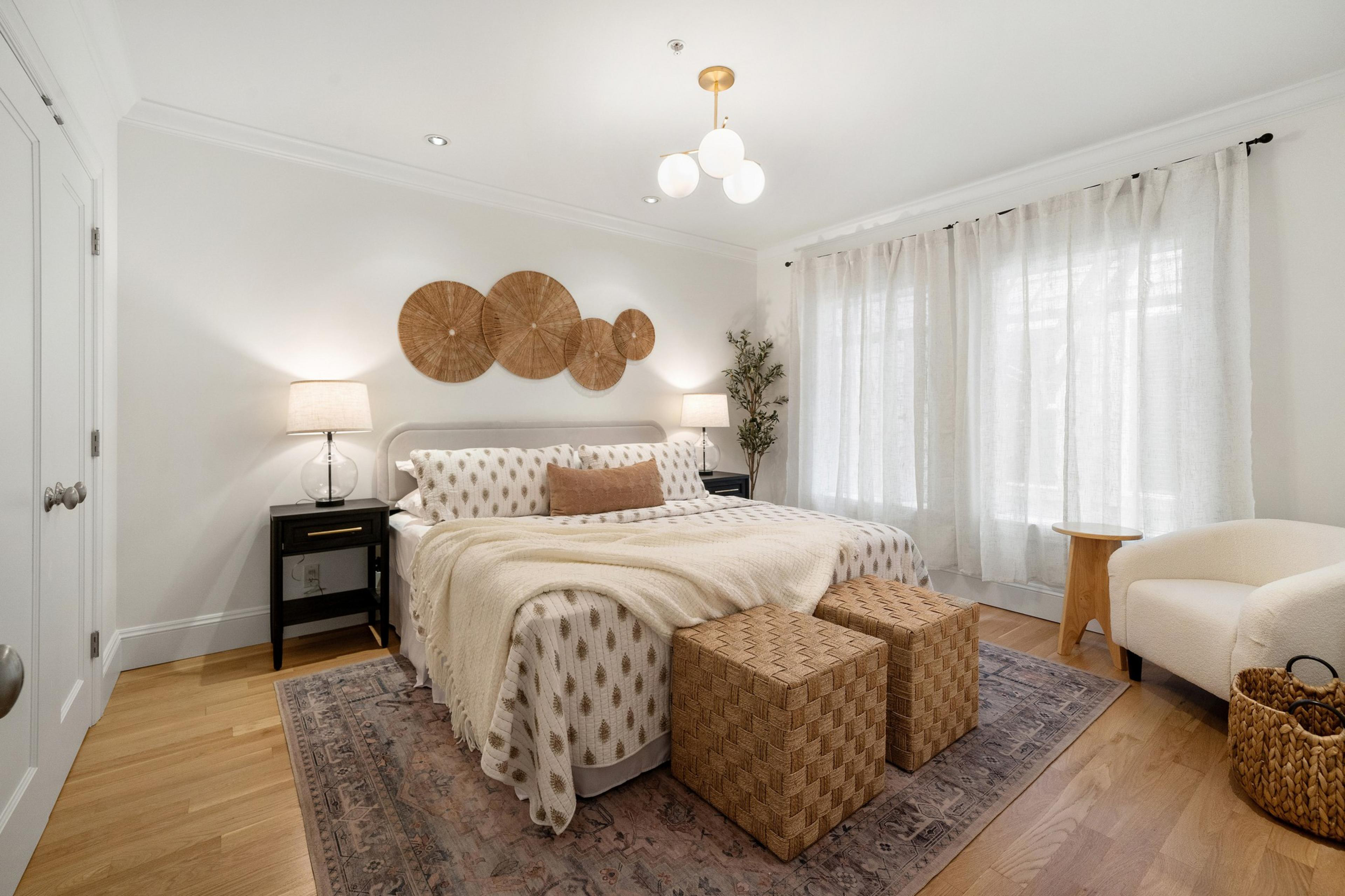 A cozy bedroom features a large bed with a patterned duvet, flanked by two nightstands with lamps. Woven wall decor, cushioned seats, and large curtained windows complete the look.