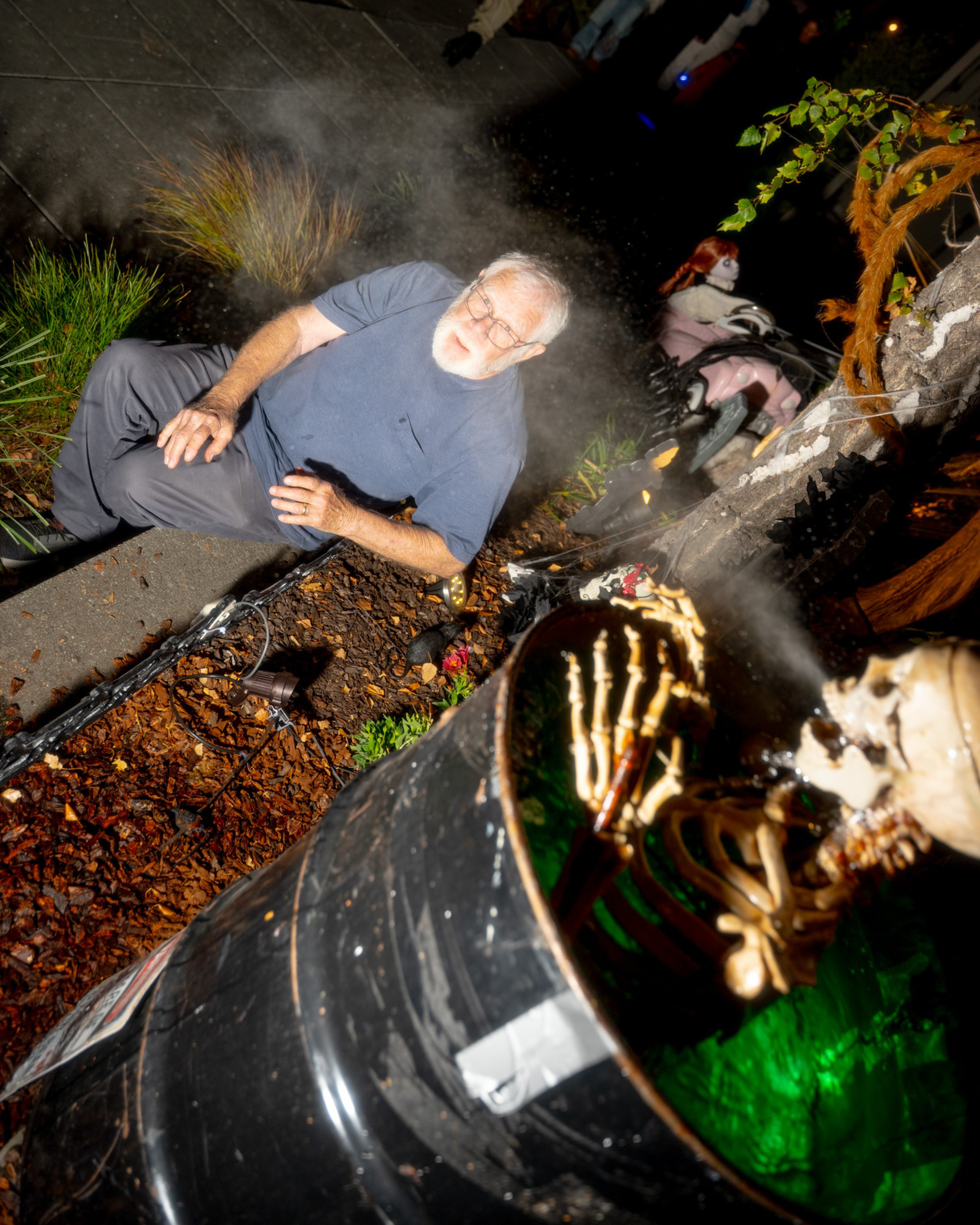 An older man with a white beard sits on the ground near a smoking barrel containing a skeleton with glowing green light inside.