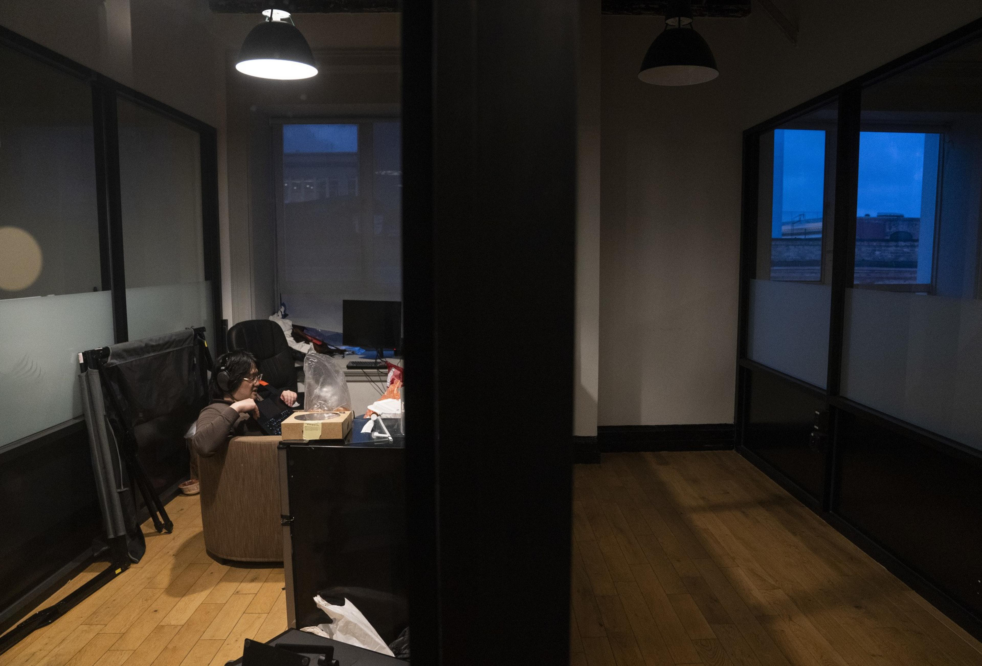 A person is sitting at a cluttered desk in a dimly lit room, wearing headphones and likely working on a computer. The room is divided by a glass wall.