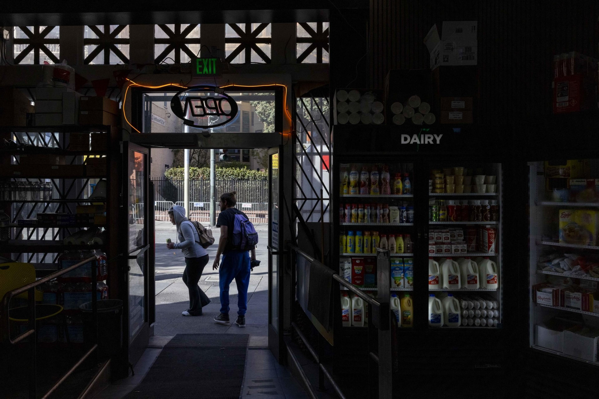 A dimly lit store interior with a glowing "Open" sign above the door. Two people exit, passing dairy shelves filled with milk and drinks.