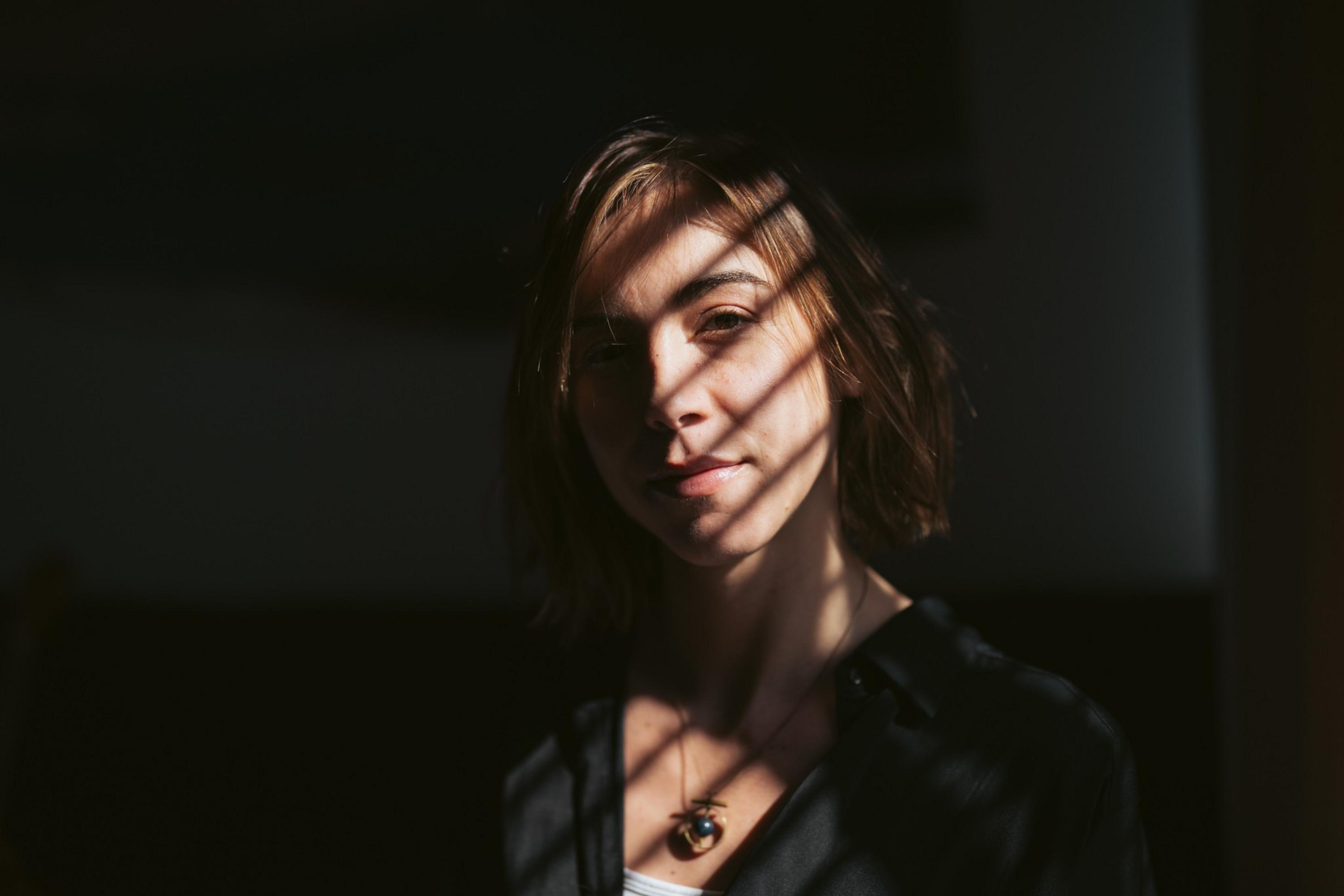 A woman with short brown hair stands in partial shadow with light and shadow patterns cast across her face and upper body, wearing a necklace and dark clothing.