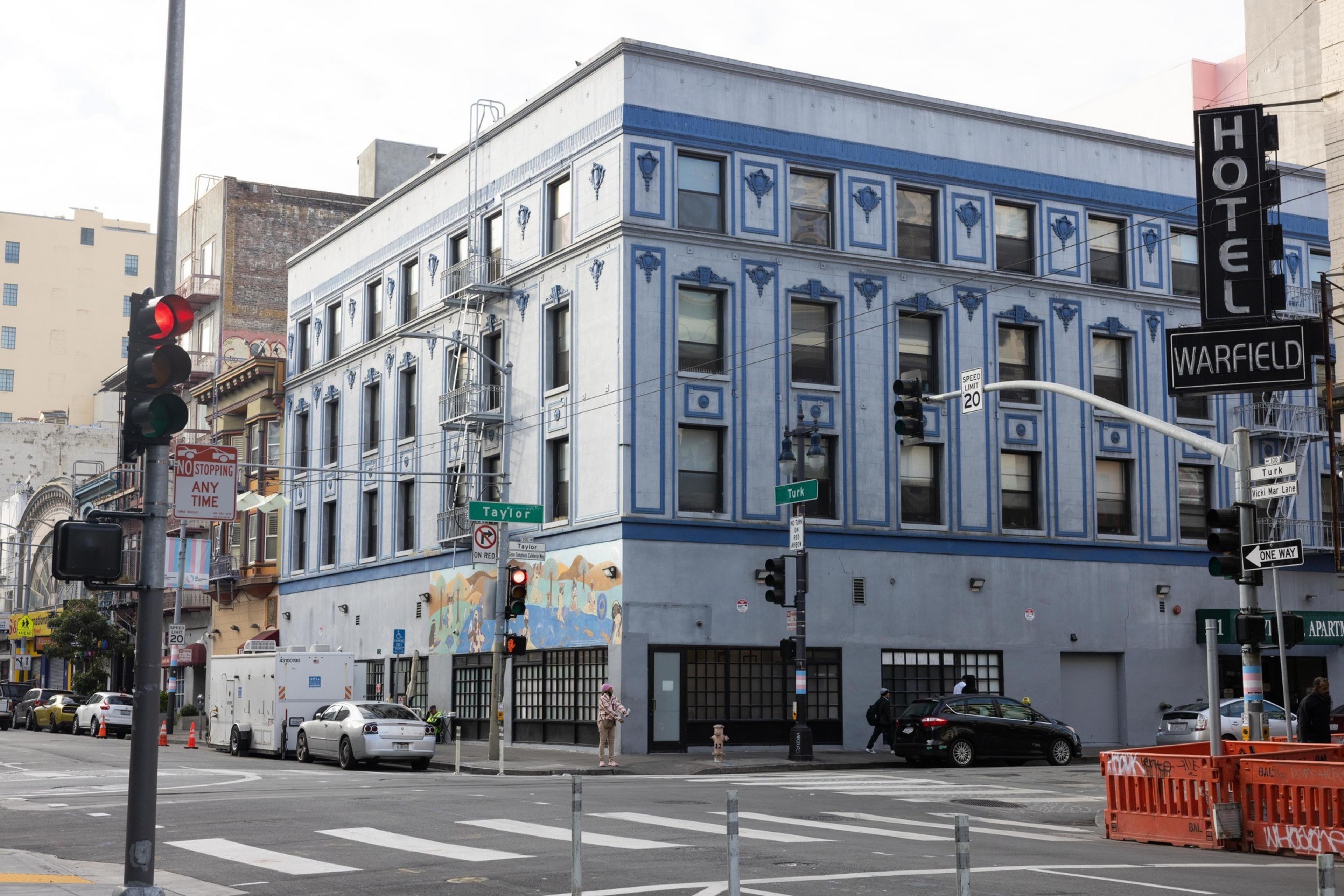 A blue, ornate, three-story building stands at an intersection. It has &quot;HOTEL WARFIELD&quot; signs, a mural, cars, and people nearby with traffic lights.