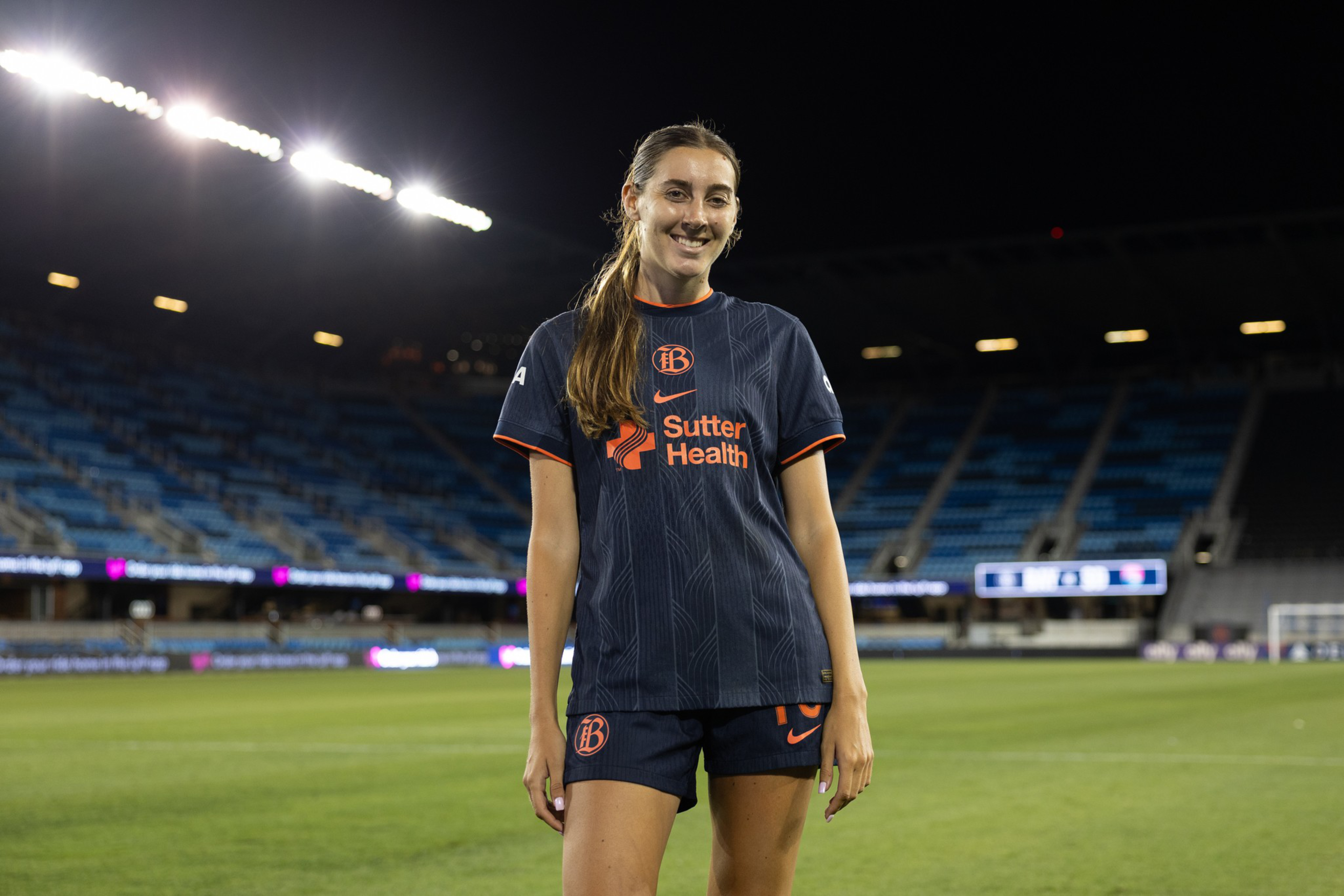 A female soccer player in a dark blue uniform with "Sutter Health" stands smiling on a lit stadium field at night, with empty blue seats in the background.