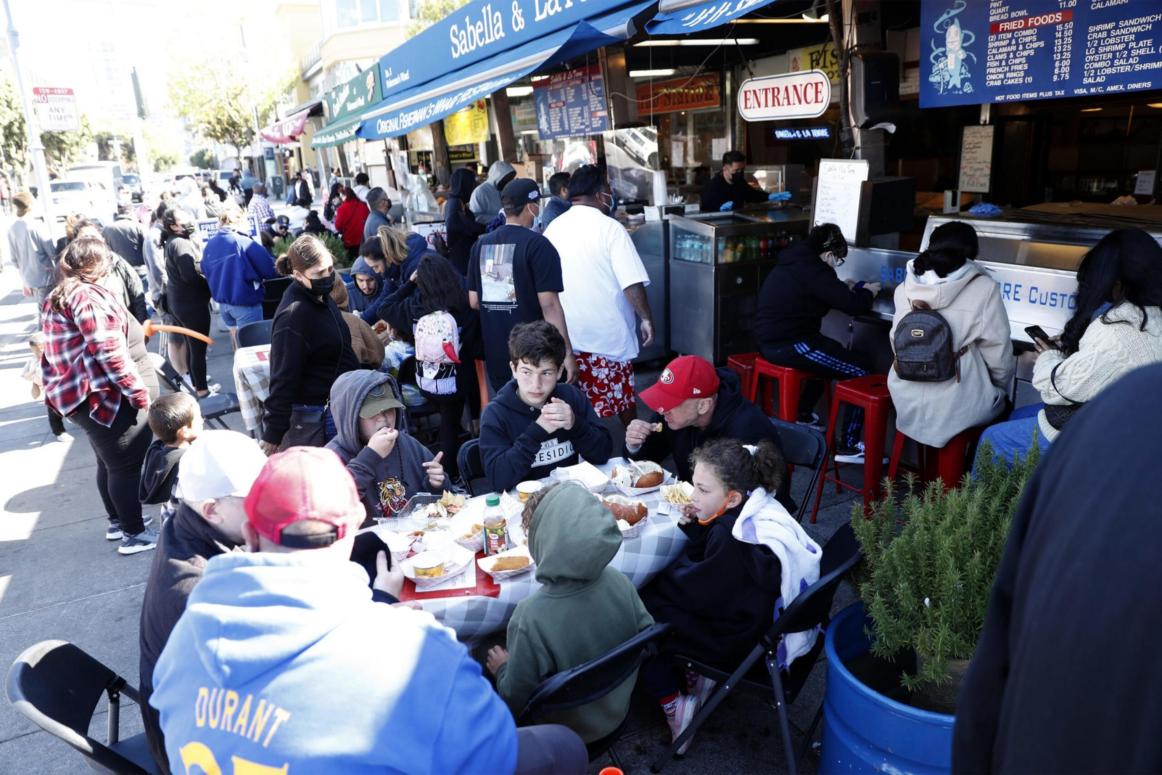 People eat and wait in line outside Sabella & La Torre seafood restaurant on a busy sunny day, with menus and signs visible above the counters.