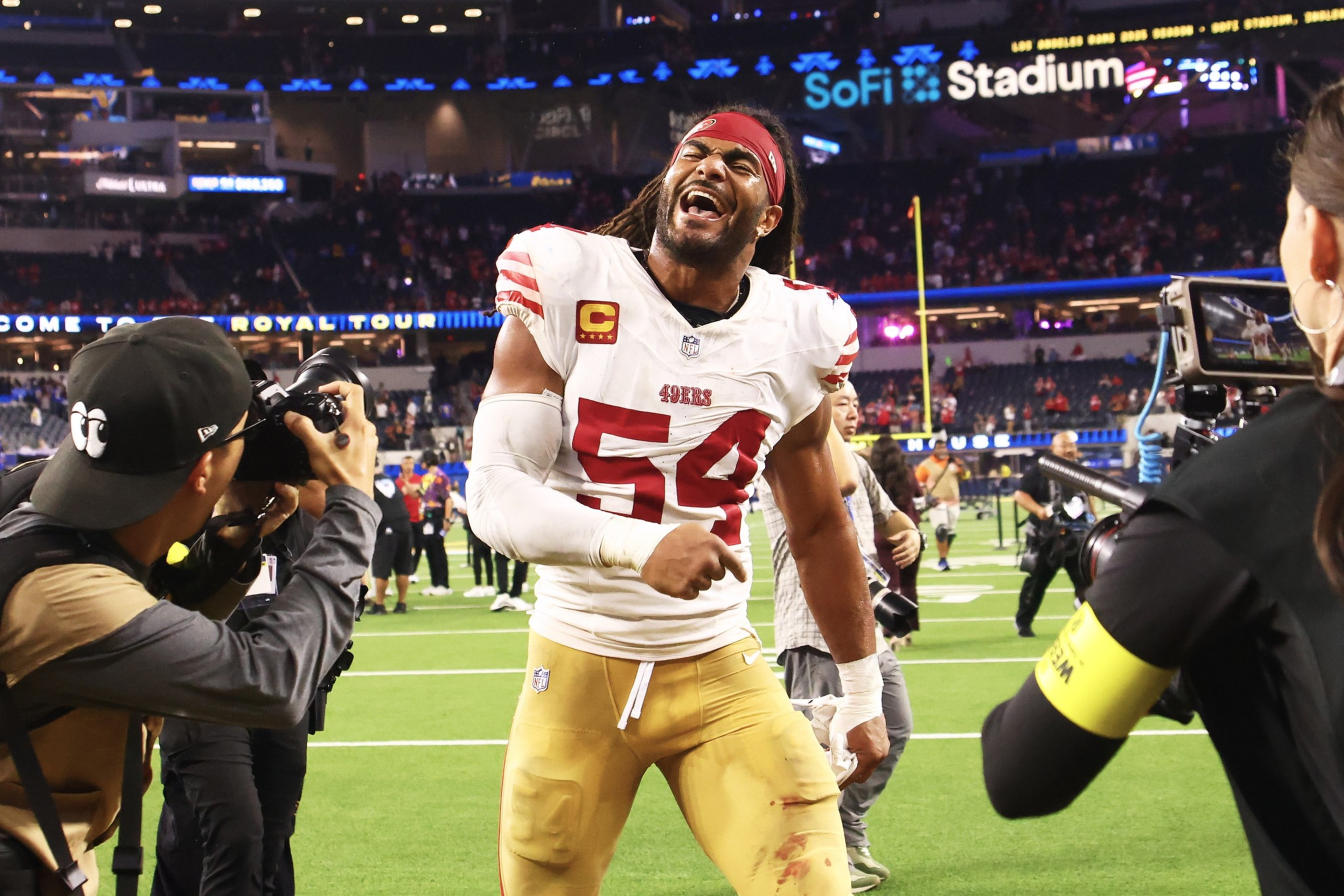 A football player in a 49ers uniform, number 54, smiles broadly and poses while photographers capture his celebration on the field.