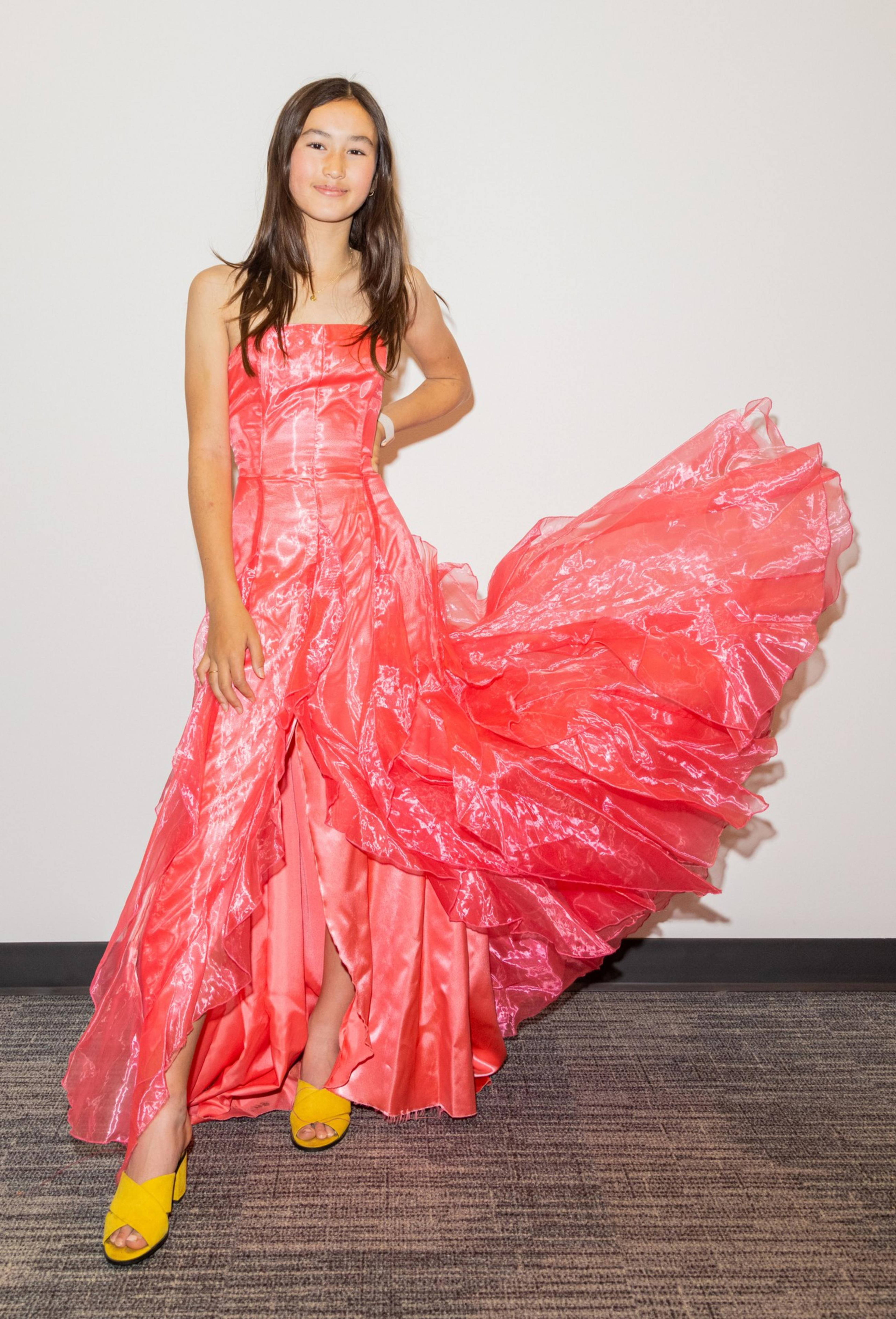 A girl stands smiling in a flowing coral dress with her hand on her hip, wearing yellow sandals.