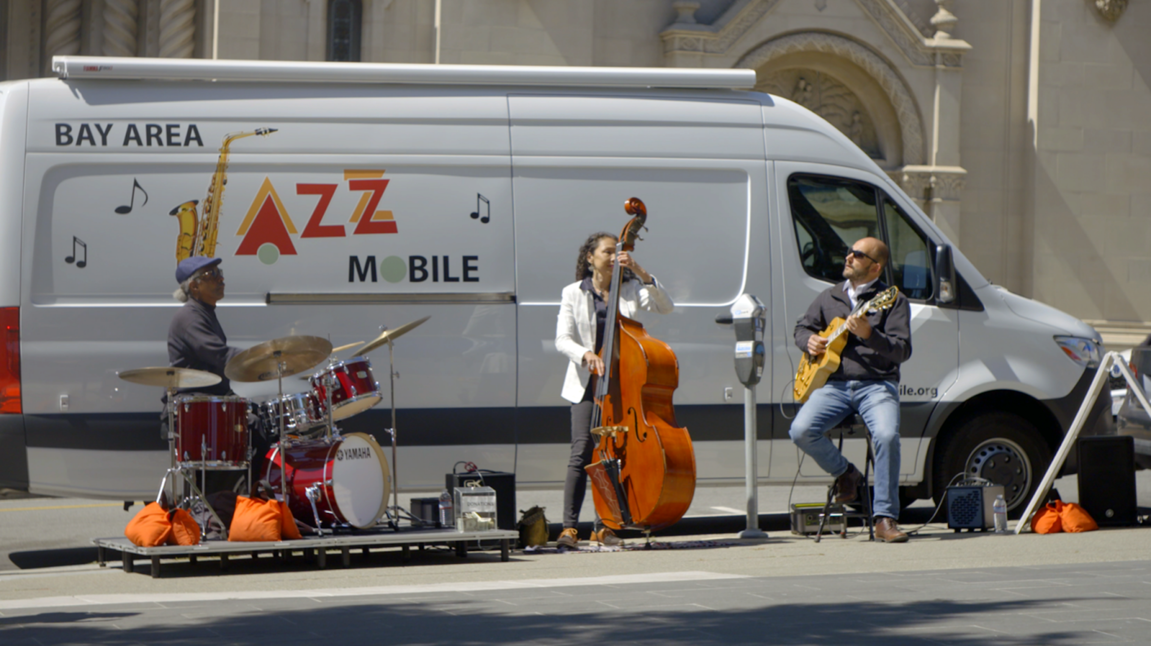 Three musicians perform outside a white van labeled "Bay Area Jazz Mobile," playing drums, upright bass, and guitar on a sunny street.