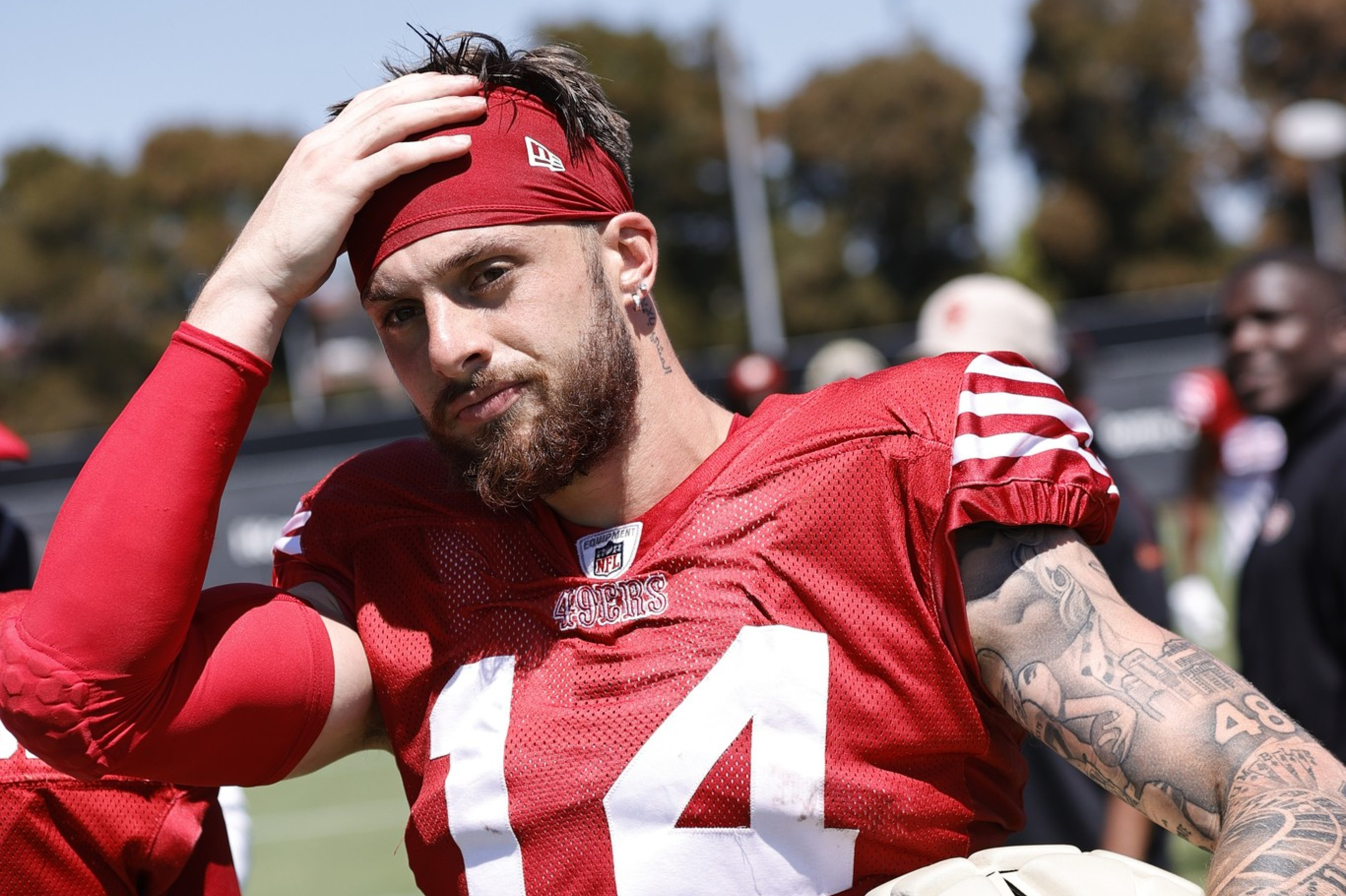 A football player in a red jersey with the number 14 holds a helmet, wipes sweat from his head, and has tattoos on his left arm, standing on a field with people in the background.