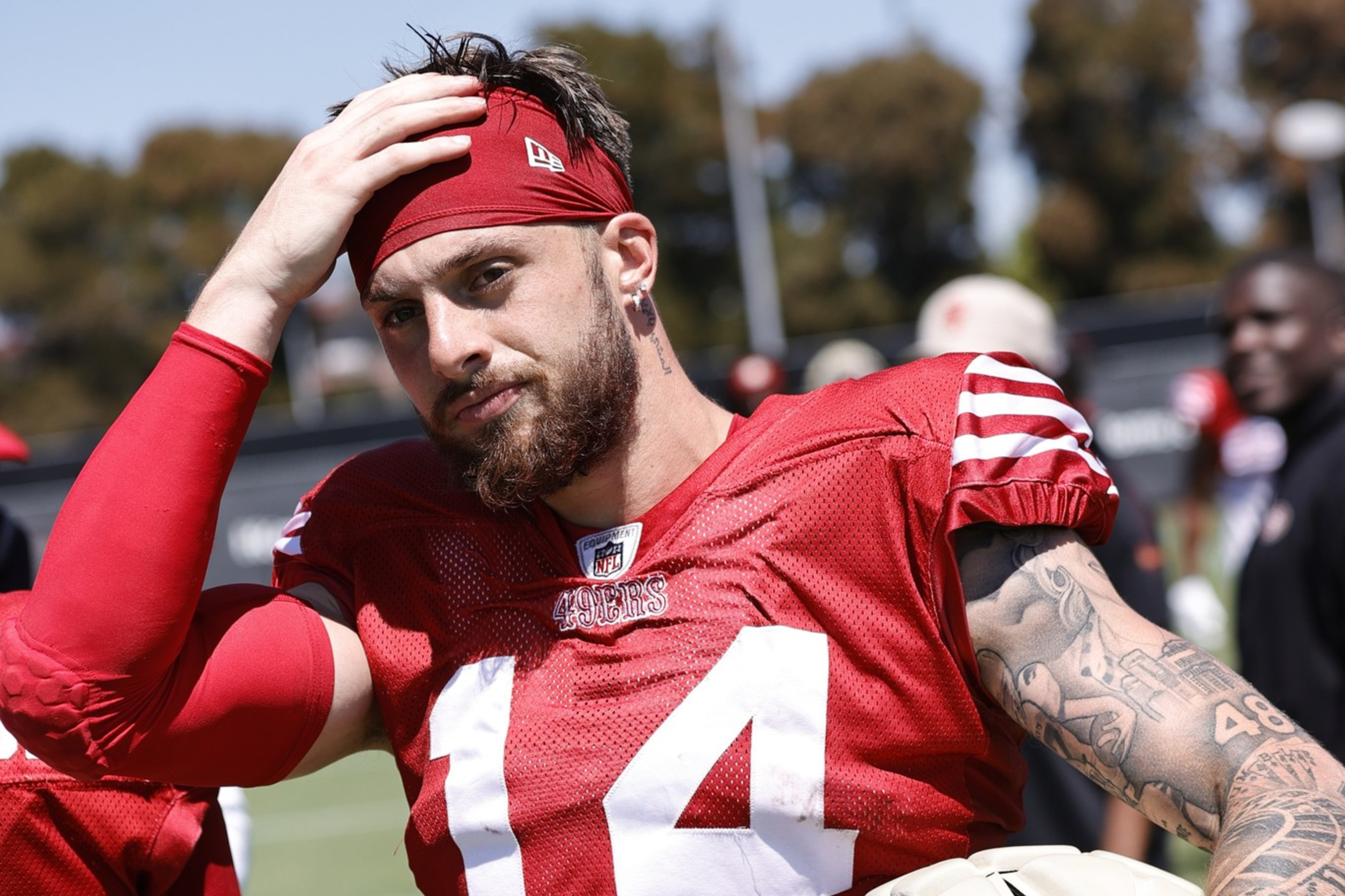 A football player in a red jersey with the number 14 holds a helmet, wipes sweat from his head, and has tattoos on his left arm, standing on a field with people in the background.