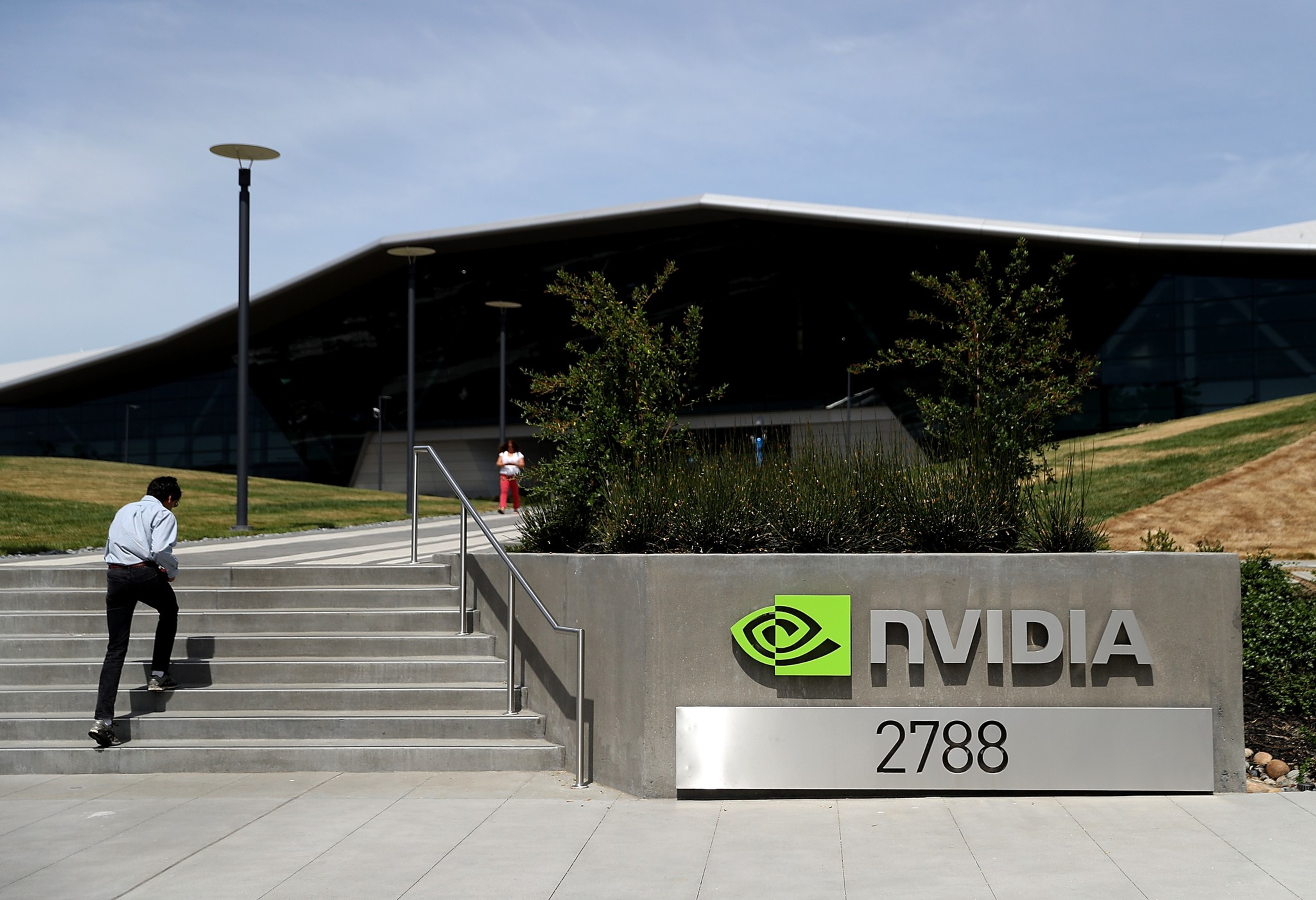 A man walks down steps at NVIDIA's modern office building entrance marked "2788" under a vibrant green logo. Another person stands in the background.