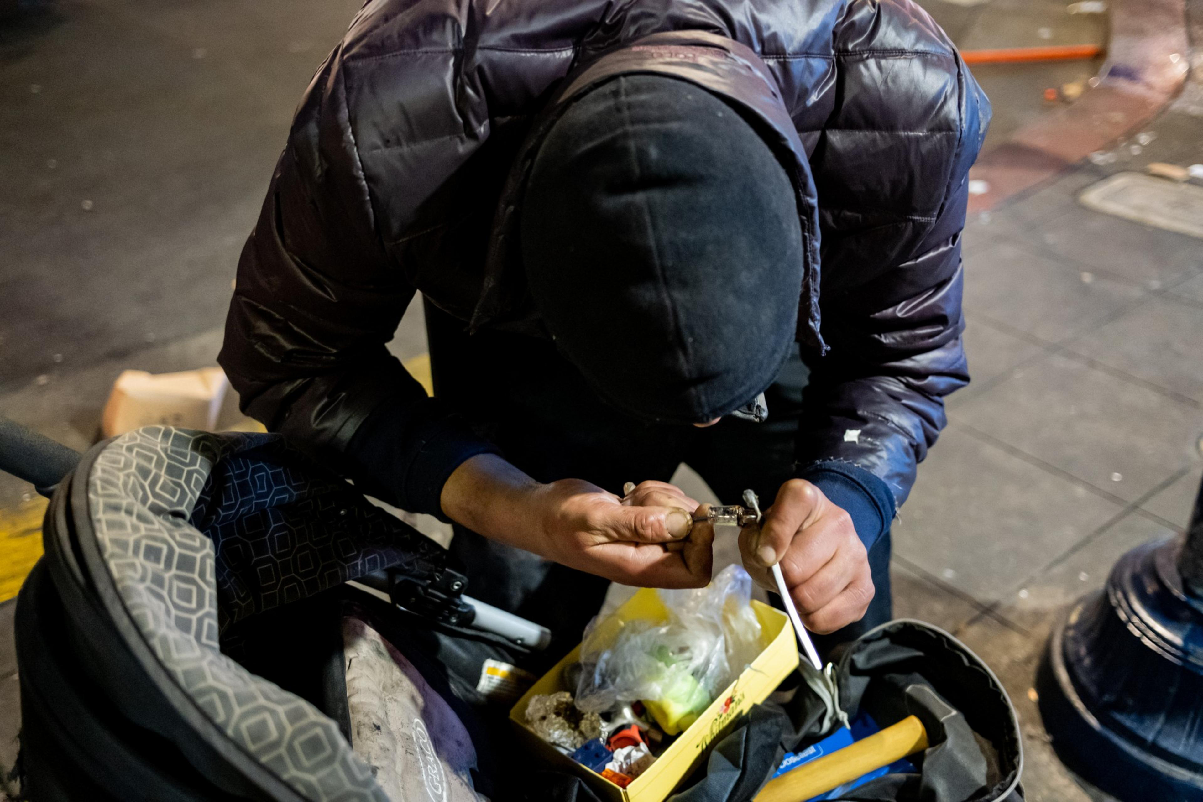 A person in a dark jacket and hat is crouched down, focused on an object in their hands, next to a stroller filled with various items, including a yellow box.