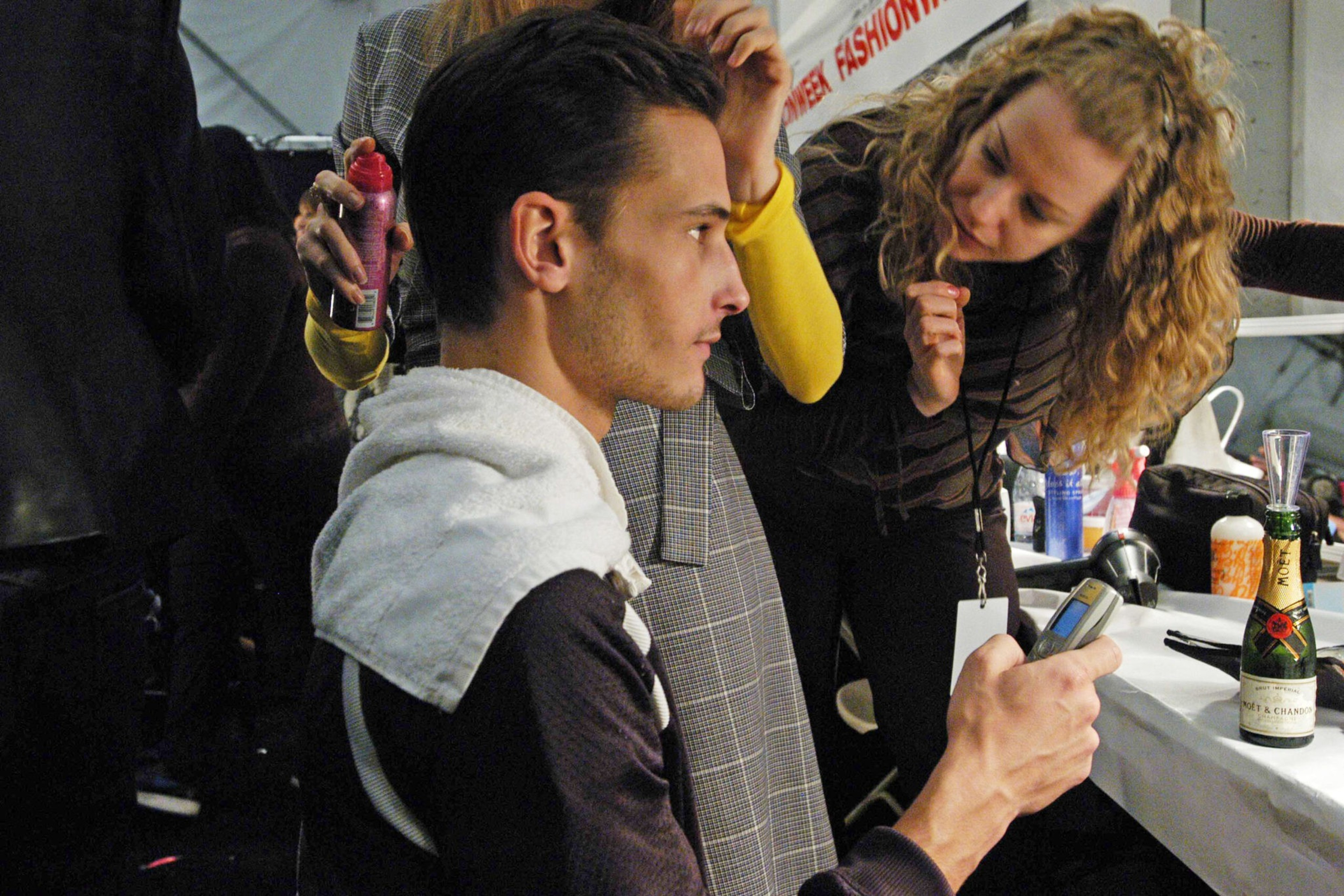 A man with a towel on his shoulders is seated while a woman styles his hair, and he holds a phone in one hand.