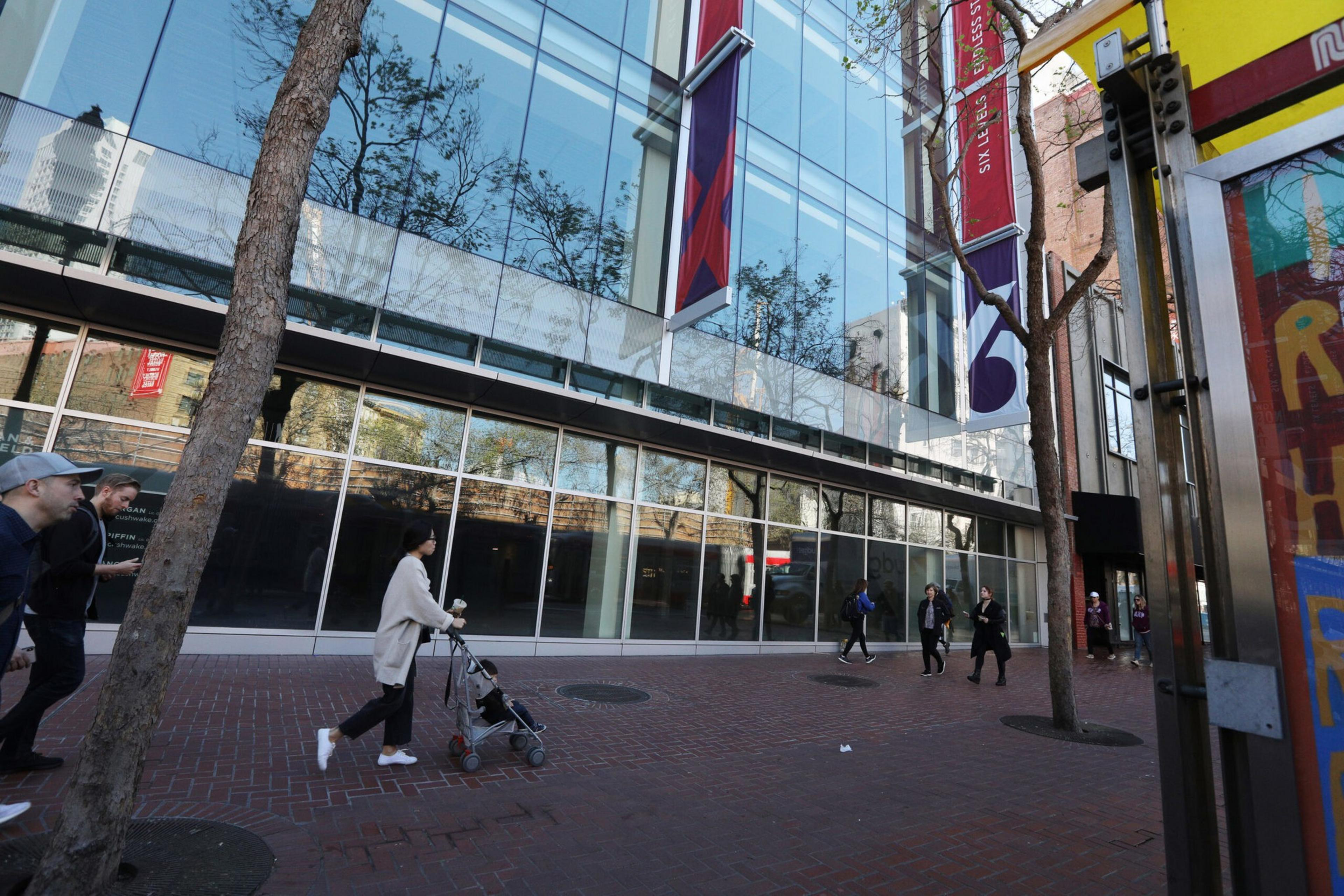 A street with several people walking past a modern glass building, some trees, and a colorful structure on the right side of the image.