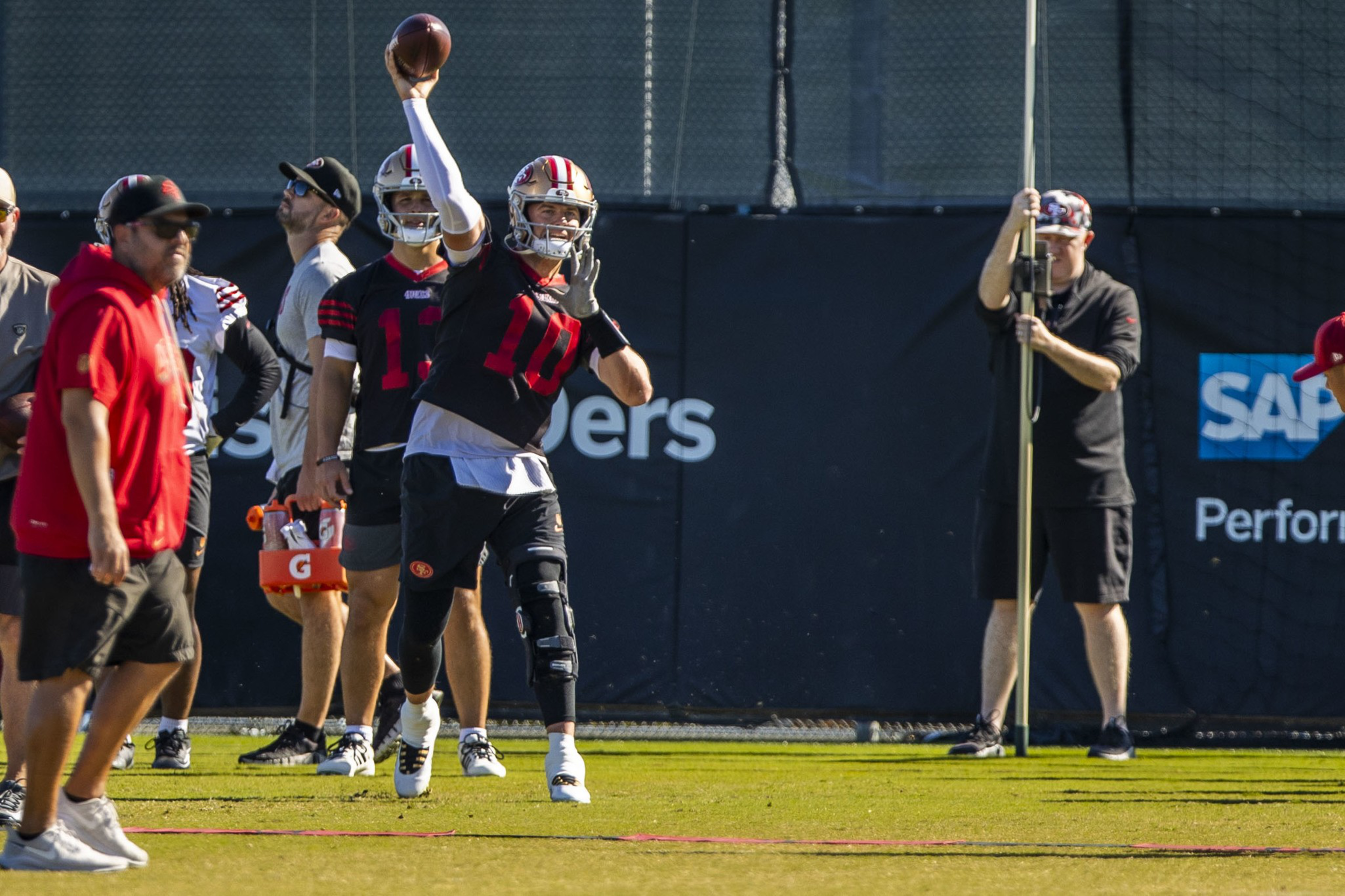 A football player in a black and red uniform throws a pass during practice while others and a cameraman watch nearby.